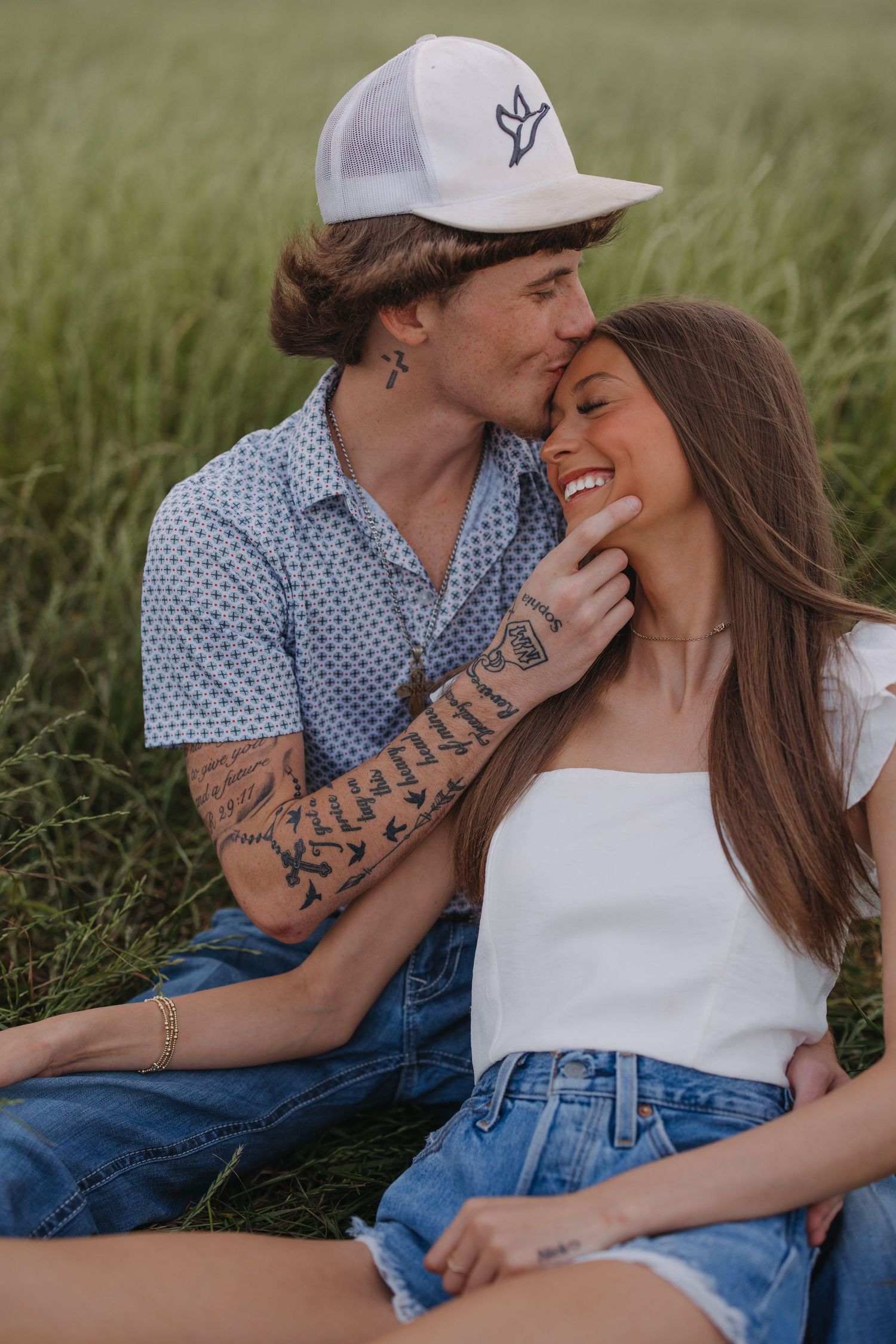 Tattooed man in a cap kisses a smiling woman sitting in a green field.