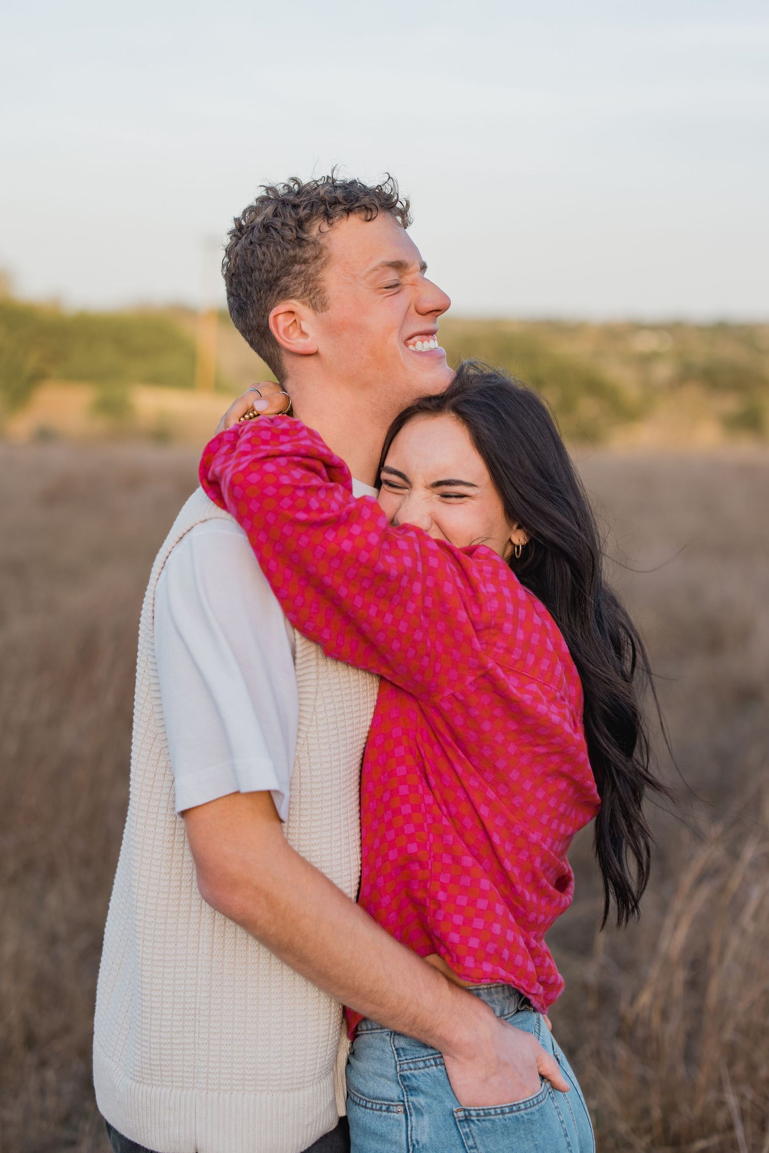 Joyful couple embracing outdoors, woman in pink top hugging man in cream vest in golden field.