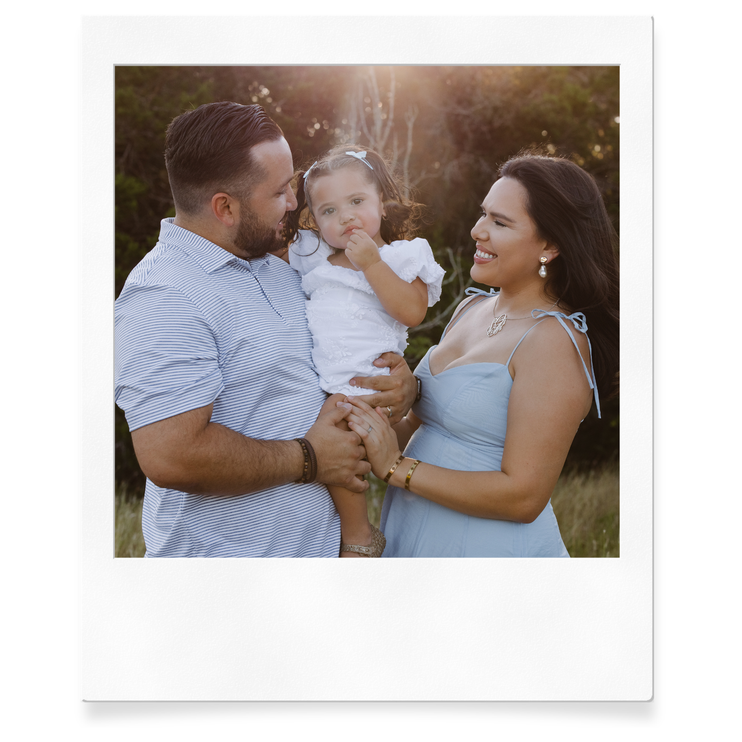Polaroid-style photo of a couple in matching blue outfits holding their smiling baby in a sunlit outdoor setting.