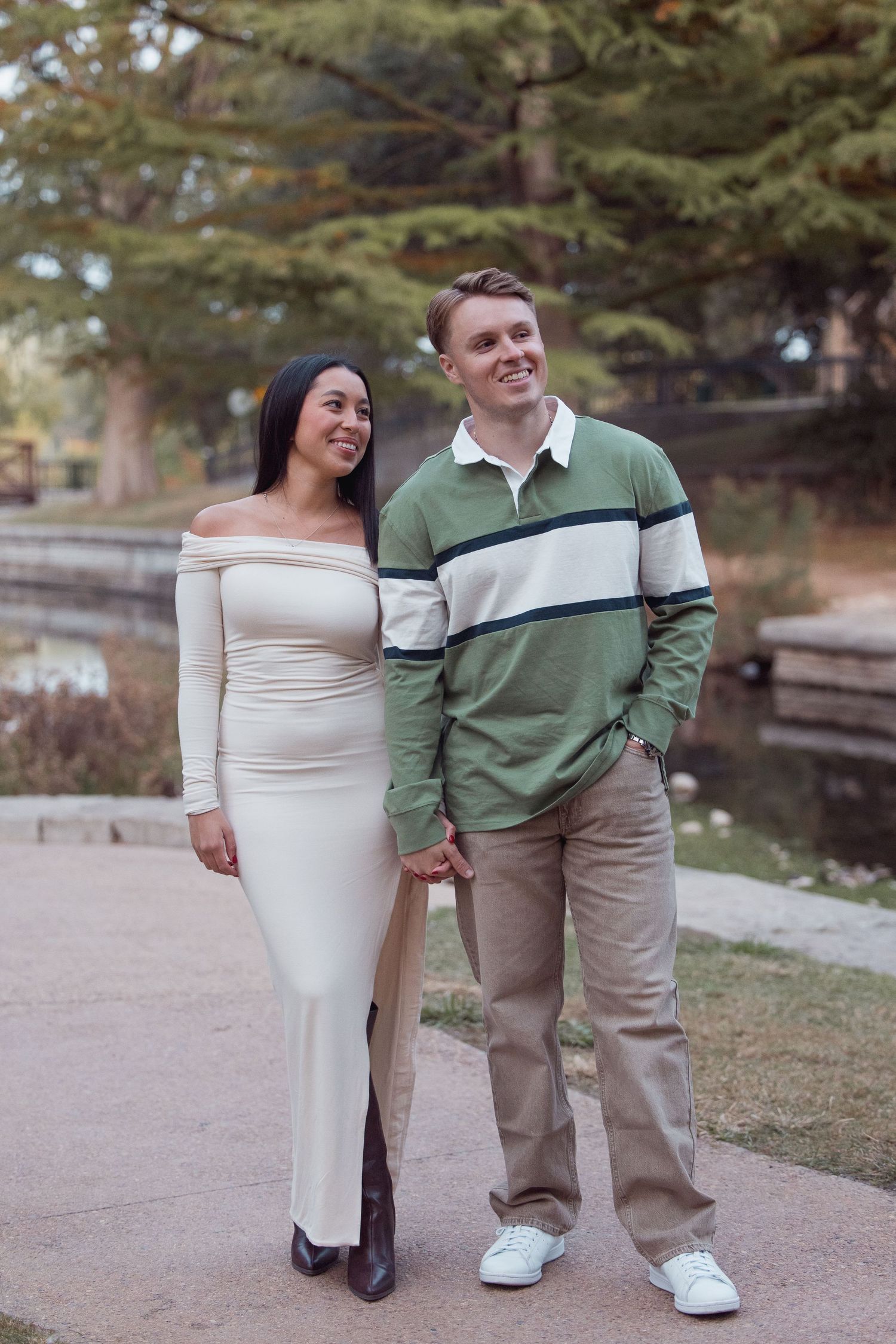 Couple standing by park pond, woman in cream off-shoulder dress and man in green striped polo shirt.