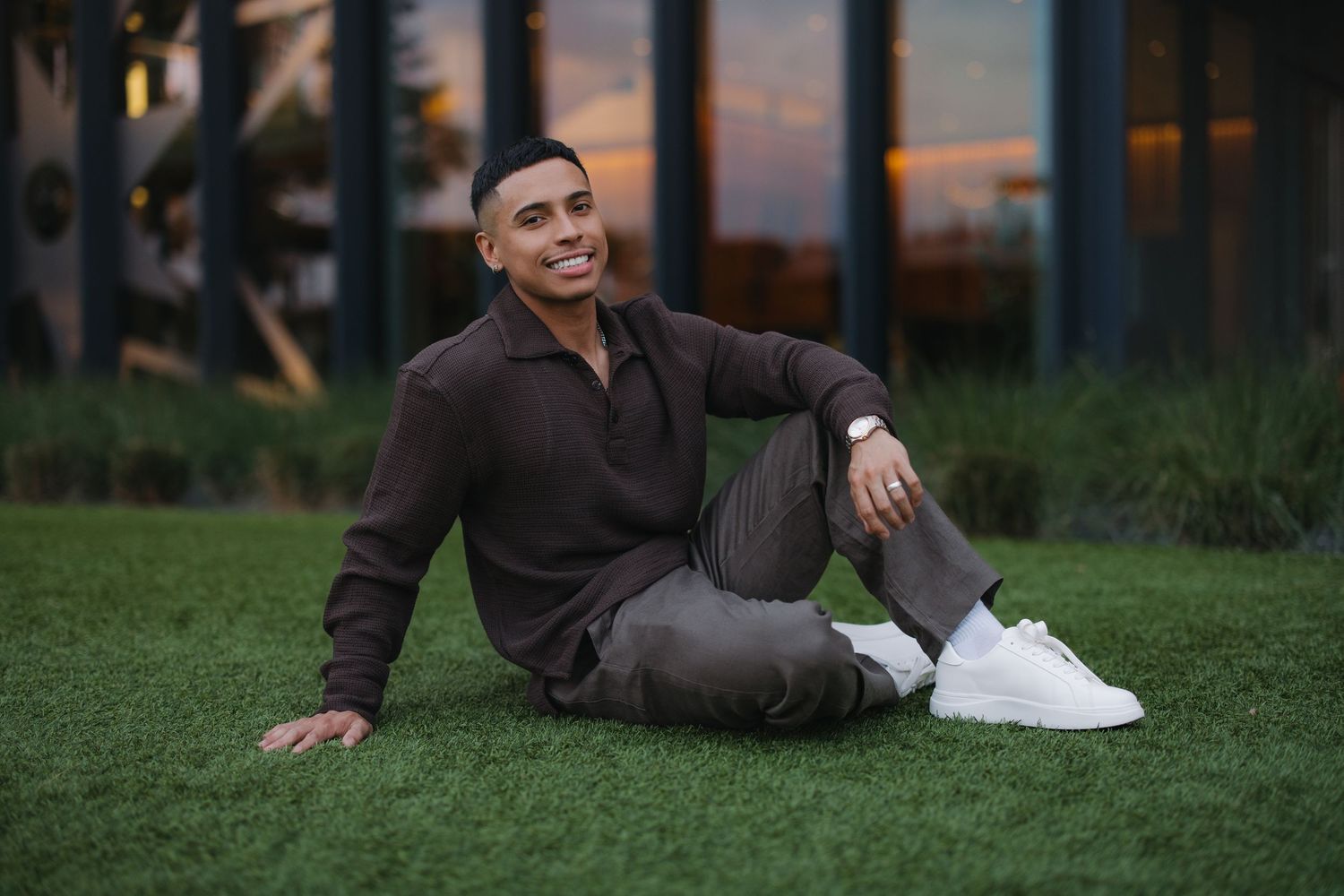 Smiling young man in brown outfit sitting relaxed on green grass at sunset outdoors.