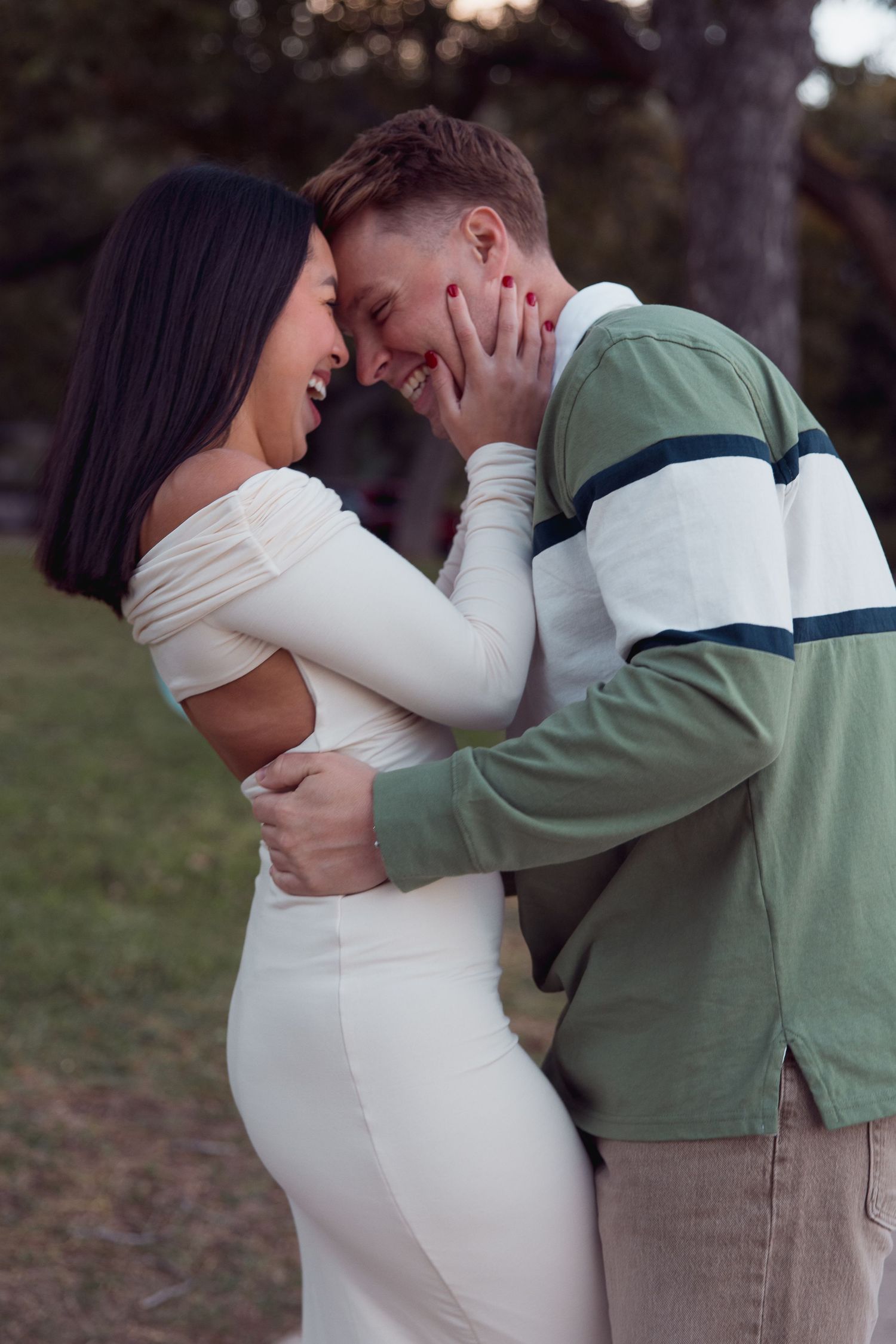 Romantic couple laughing and touching foreheads outdoors, woman in white dress and man in green shirt.