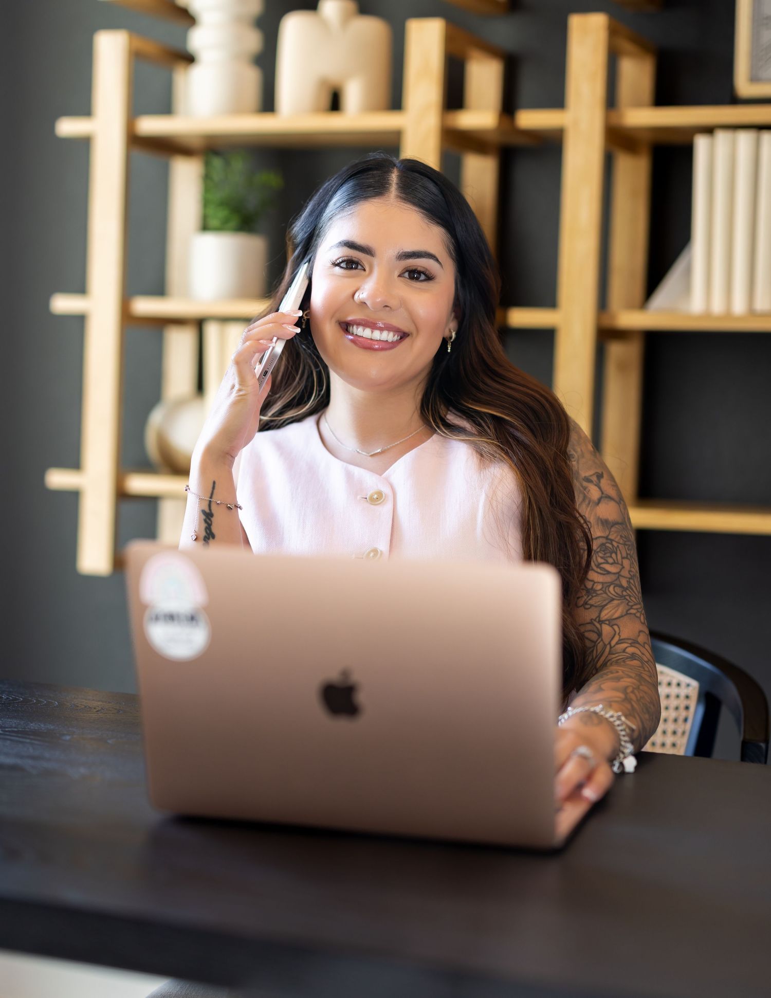 Smiling tattooed woman talking on phone while working on rose gold MacBook laptop at desk.