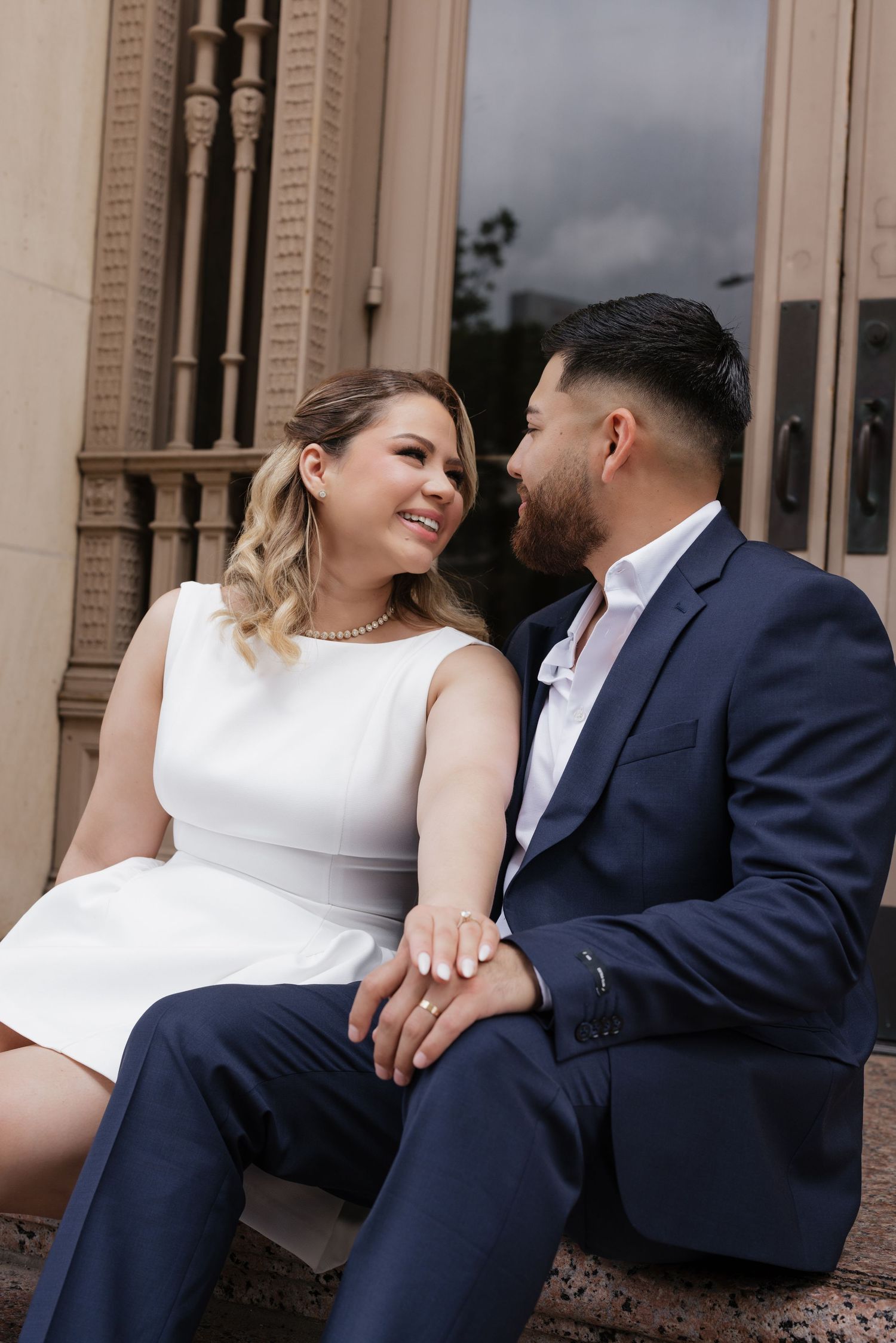 Couple sitting on steps of elegant building, woman in white dress smiling at man in navy suit holding hands.