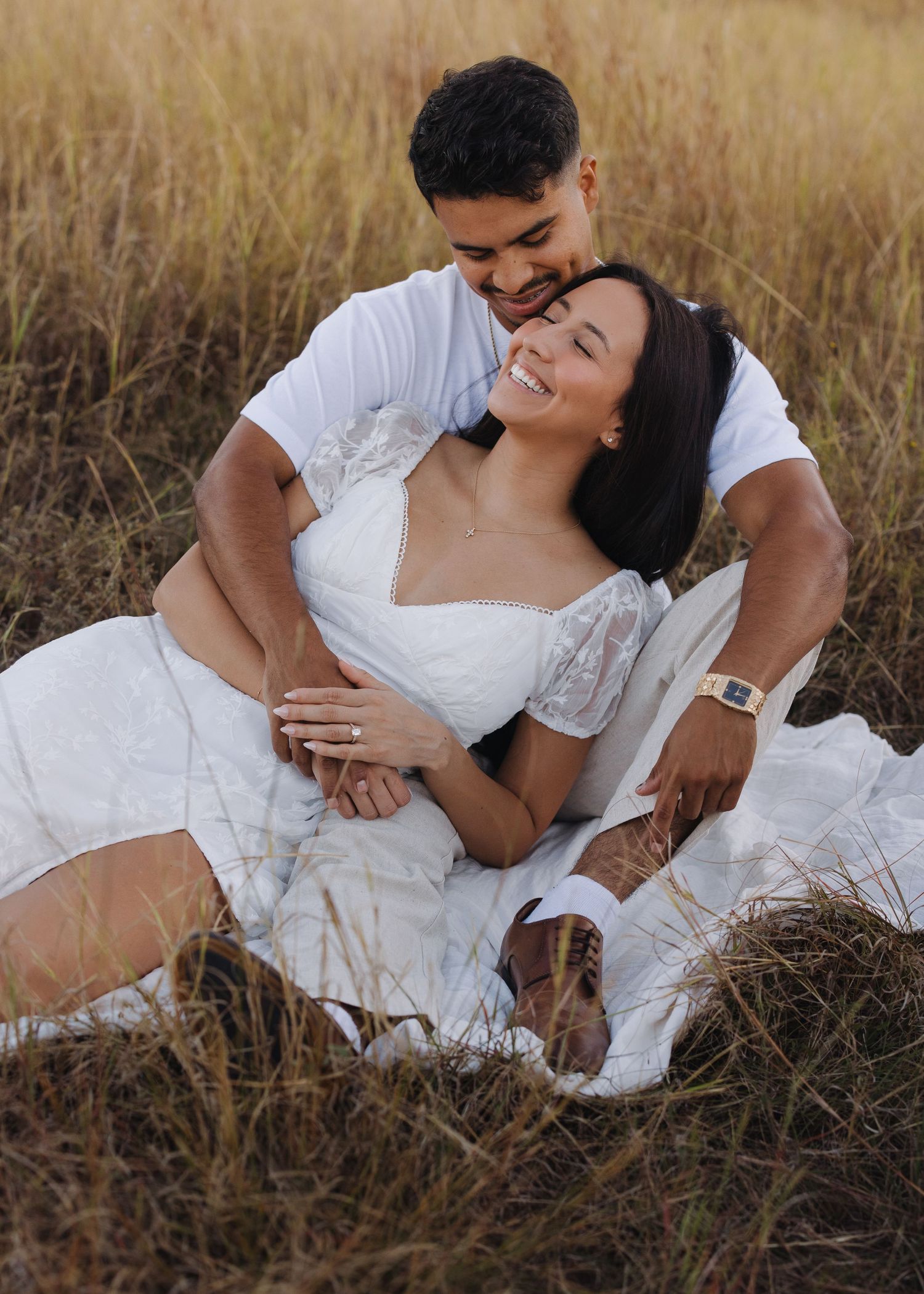 Happy couple in white outfits sitting in golden grass field during romantic outdoor photo session.