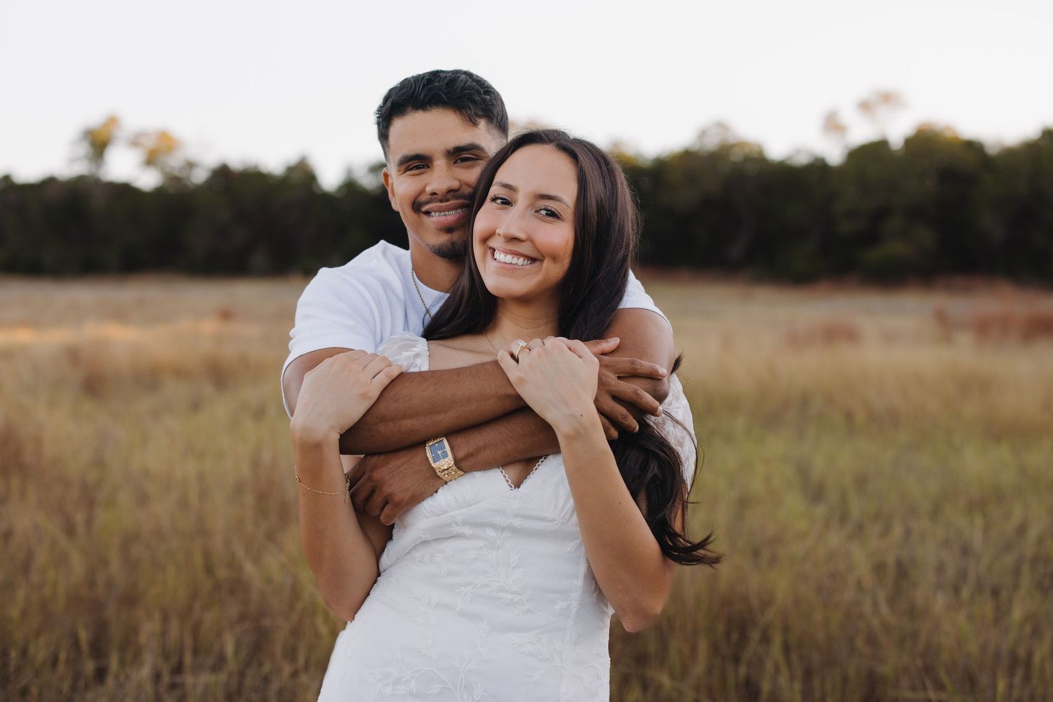 Happy couple embracing in a golden field, woman in white dress smiling as man hugs her from behind.