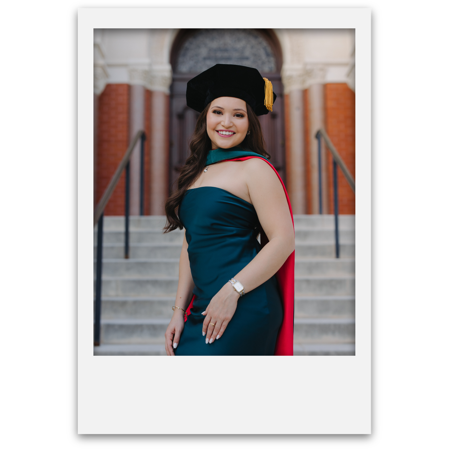 Female doctoral graduate in teal strapless gown and red hood smiles confidently on university building steps.