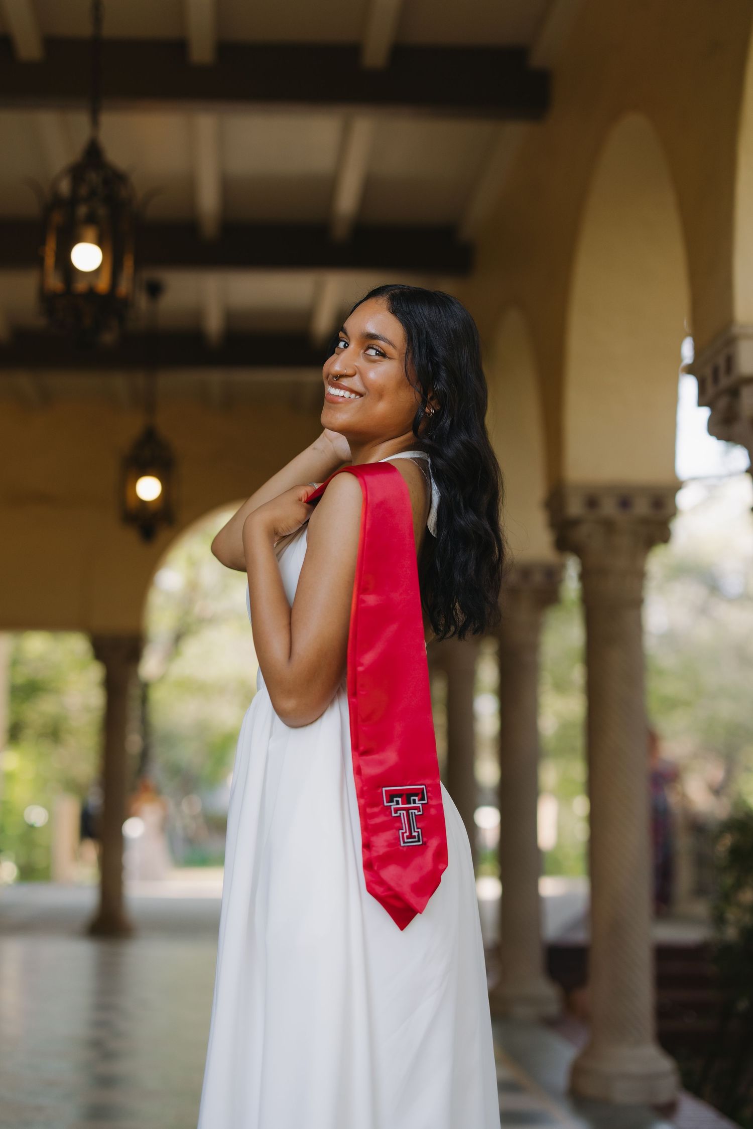 Smiling young woman in white dress with red Texas Tech graduation stole poses under elegant arched colonnade.