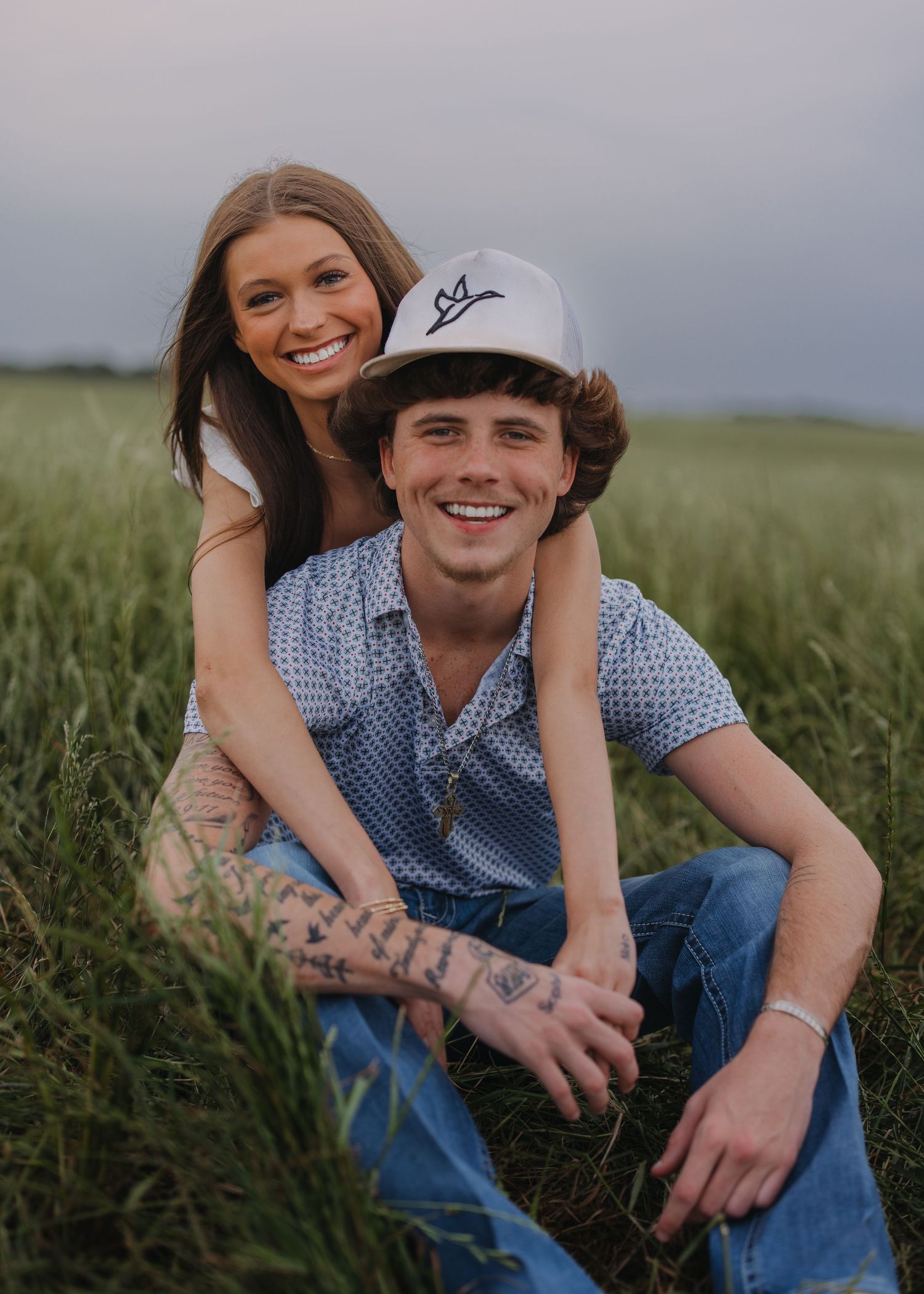 Smiling couple posing in a lush green field, man with tattoos wearing a white cap.