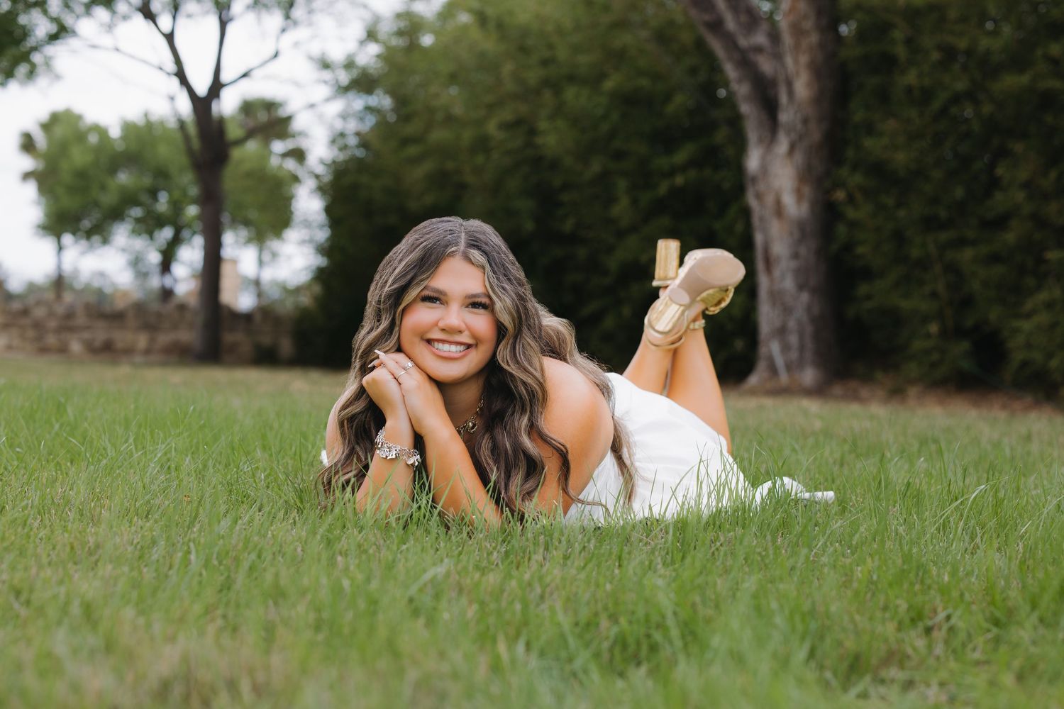 Smiling senior girl in white dress lying in green grass for outdoor portraits.