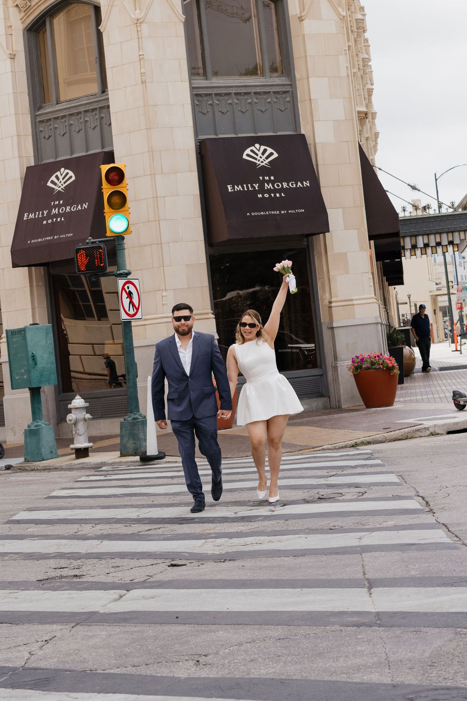 Joyful bride in white dress raises bouquet while crossing street with groom near Emily Morgan Hotel.