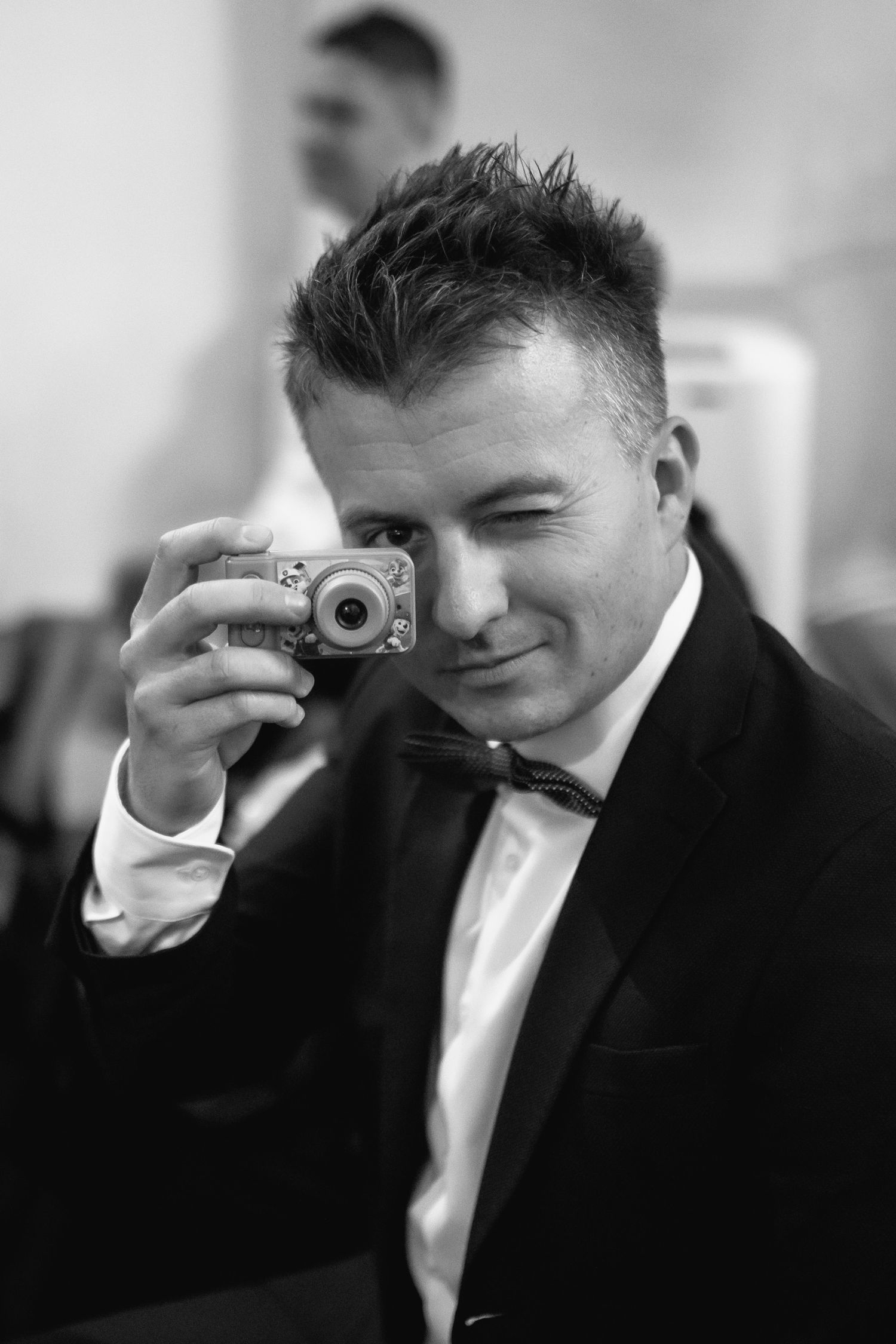 Black and white portrait of someone in a tuxedo holding a small silver digital camera with a winking expression.
