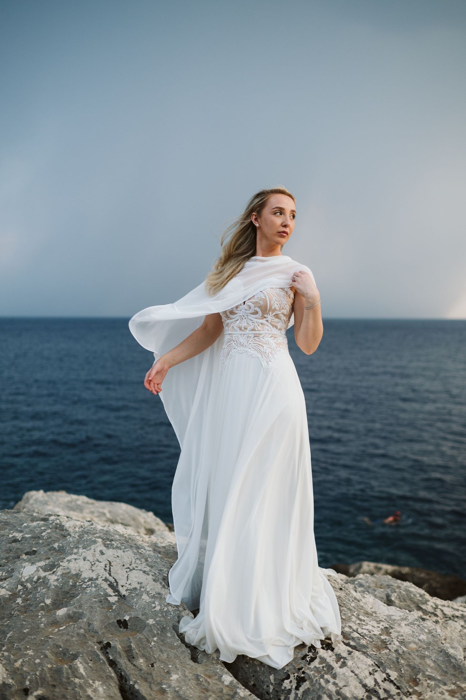 A bride in a flowing white dress stands gracefully on coastal rocks against a moody ocean backdrop.