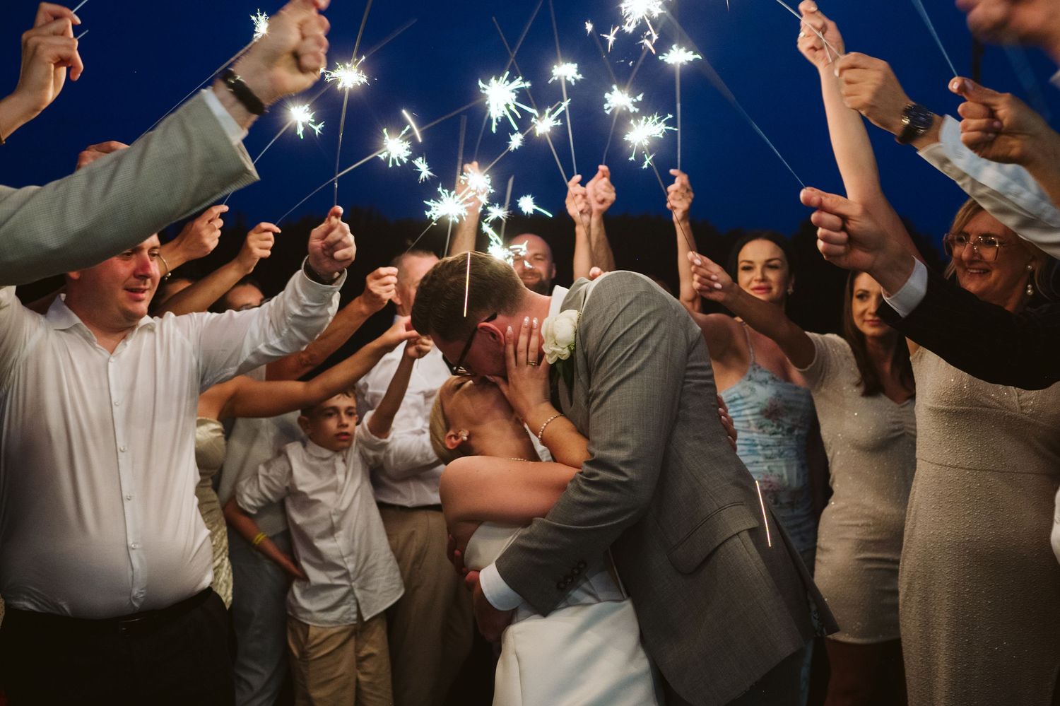 Wedding guests hold sparklers forming a magical tunnel as couple shares romantic moment during evening celebration.