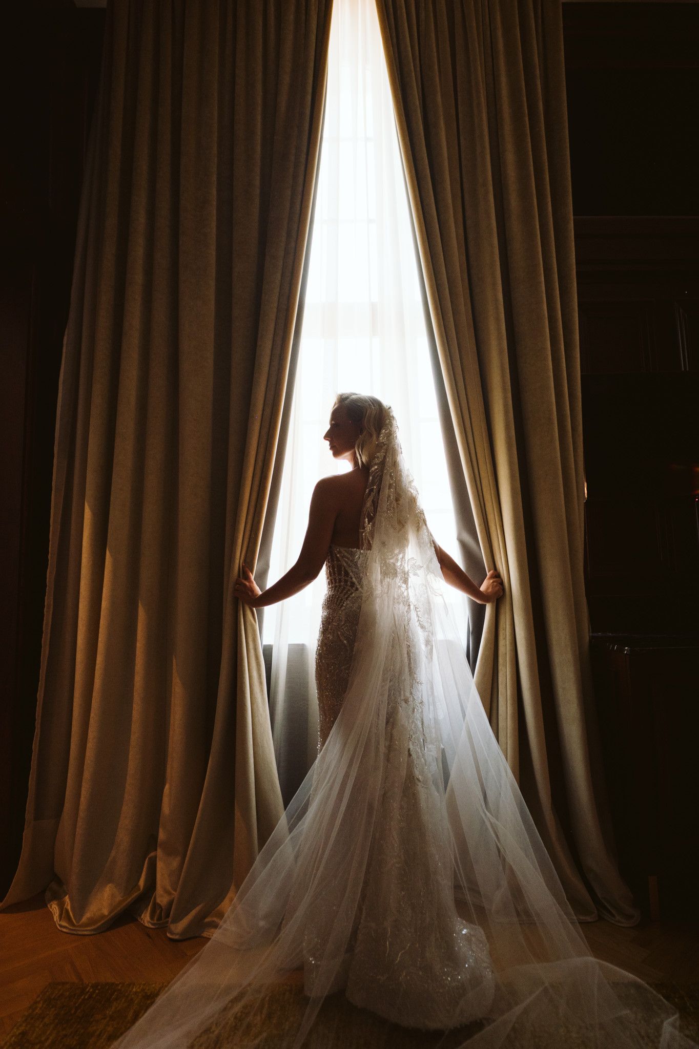 Silhouette of bride in lace gown standing between floor-length curtains by window.