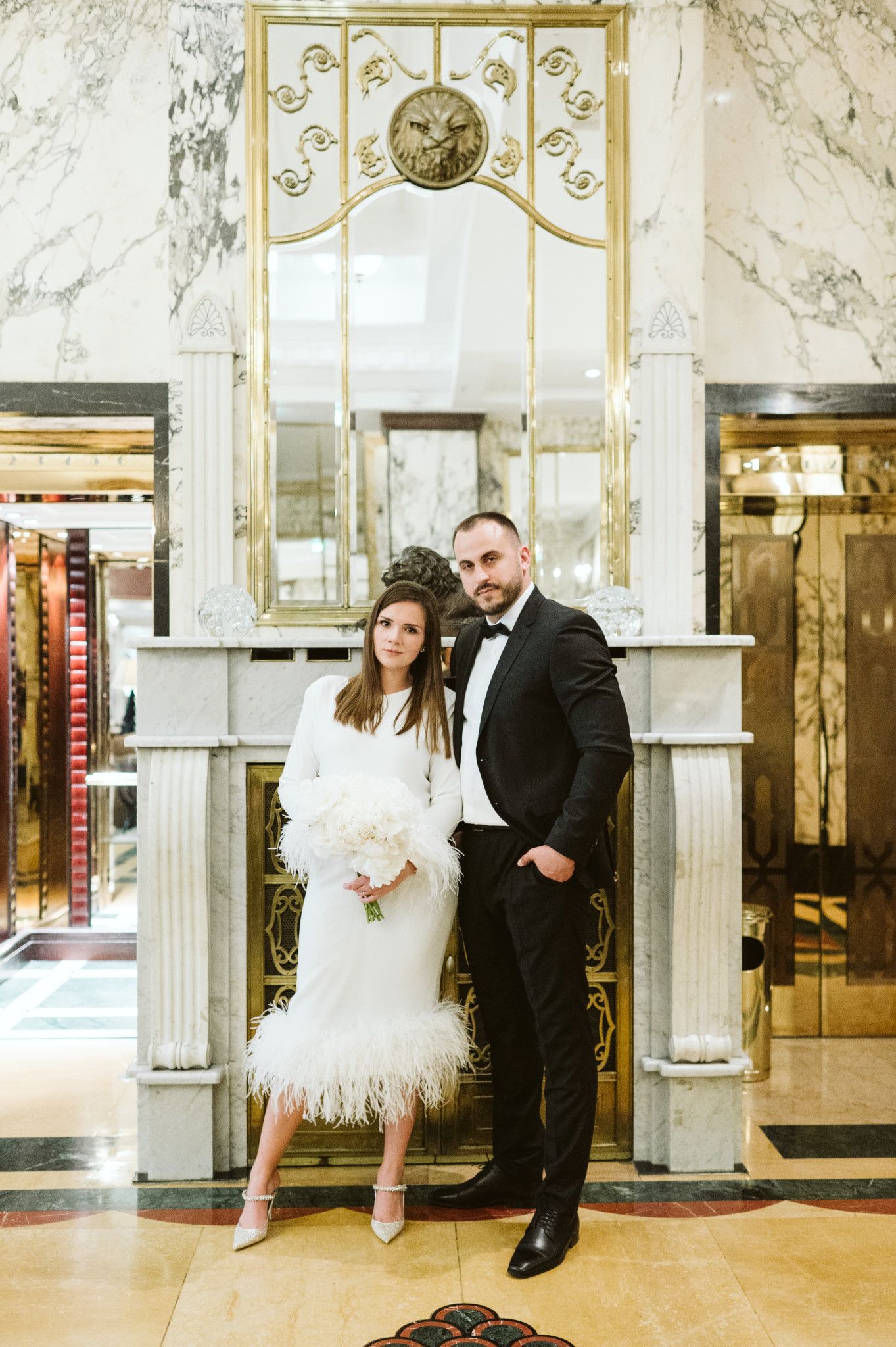 Glamorous couple poses in an ornate marble lobby with gold accents.