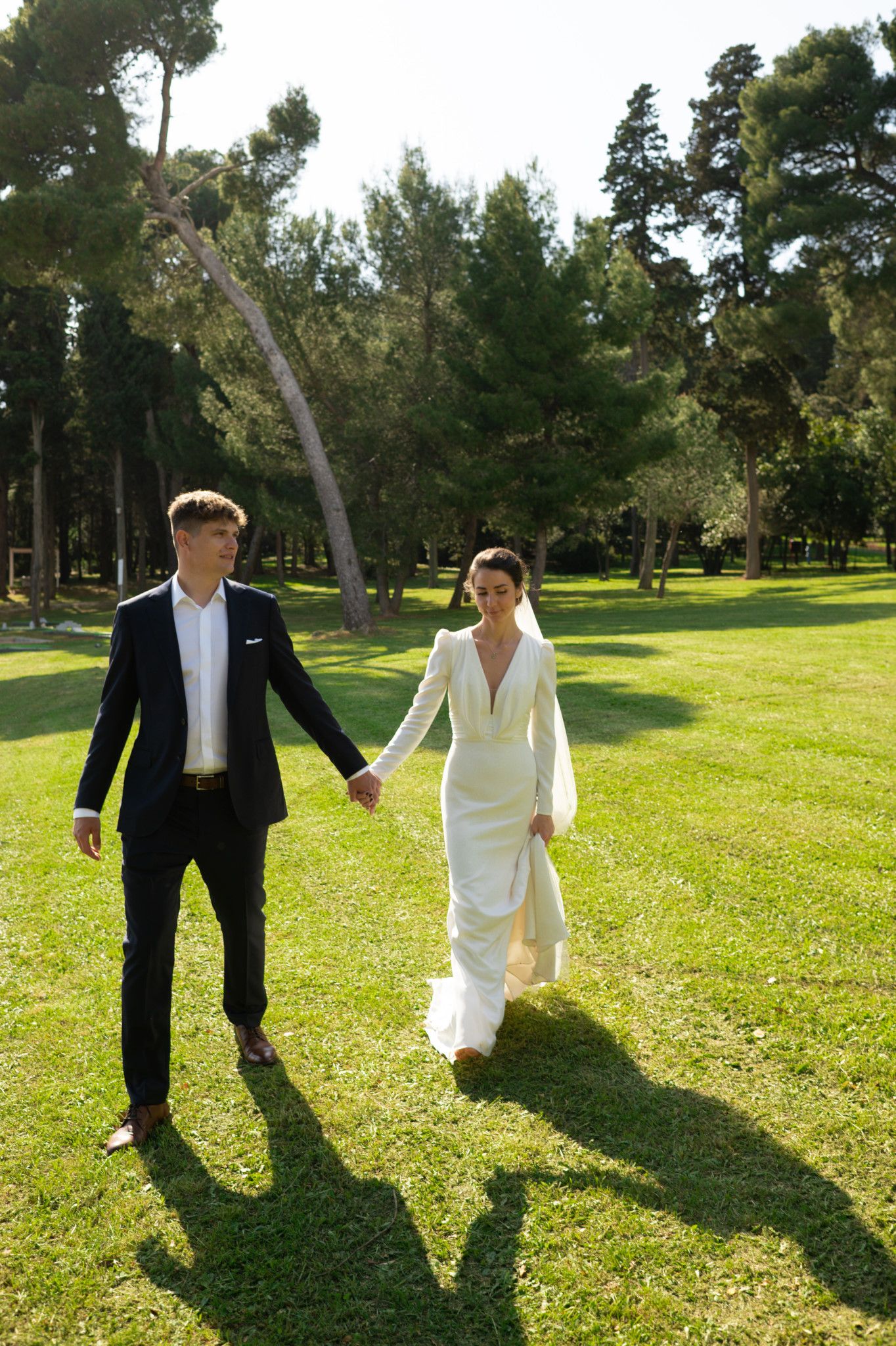 A couple in wedding attire walking hand in hand on a sunlit lawn surrounded by tall pine trees.