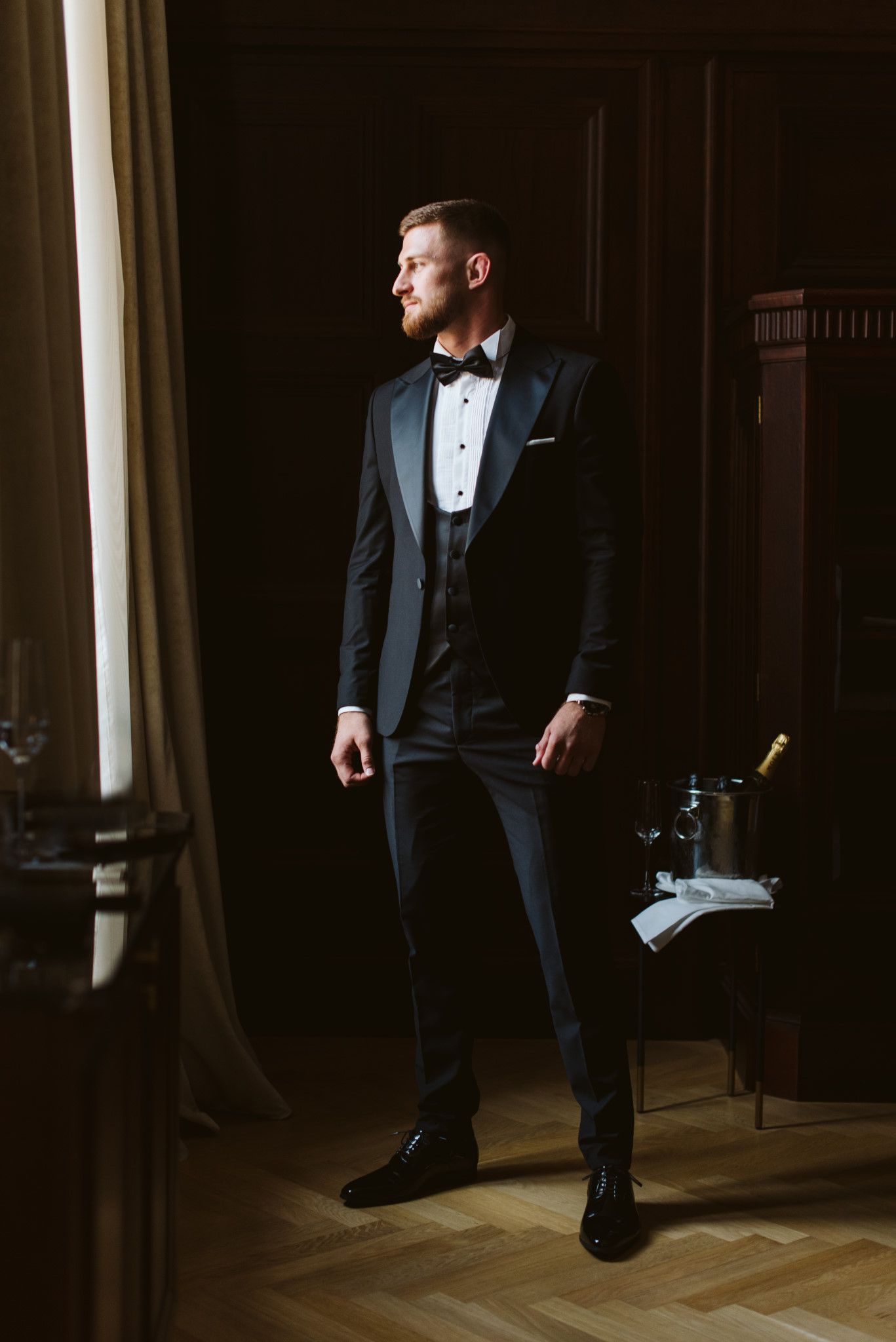 Groom in classic black tuxedo poses by window in elegant room setting.