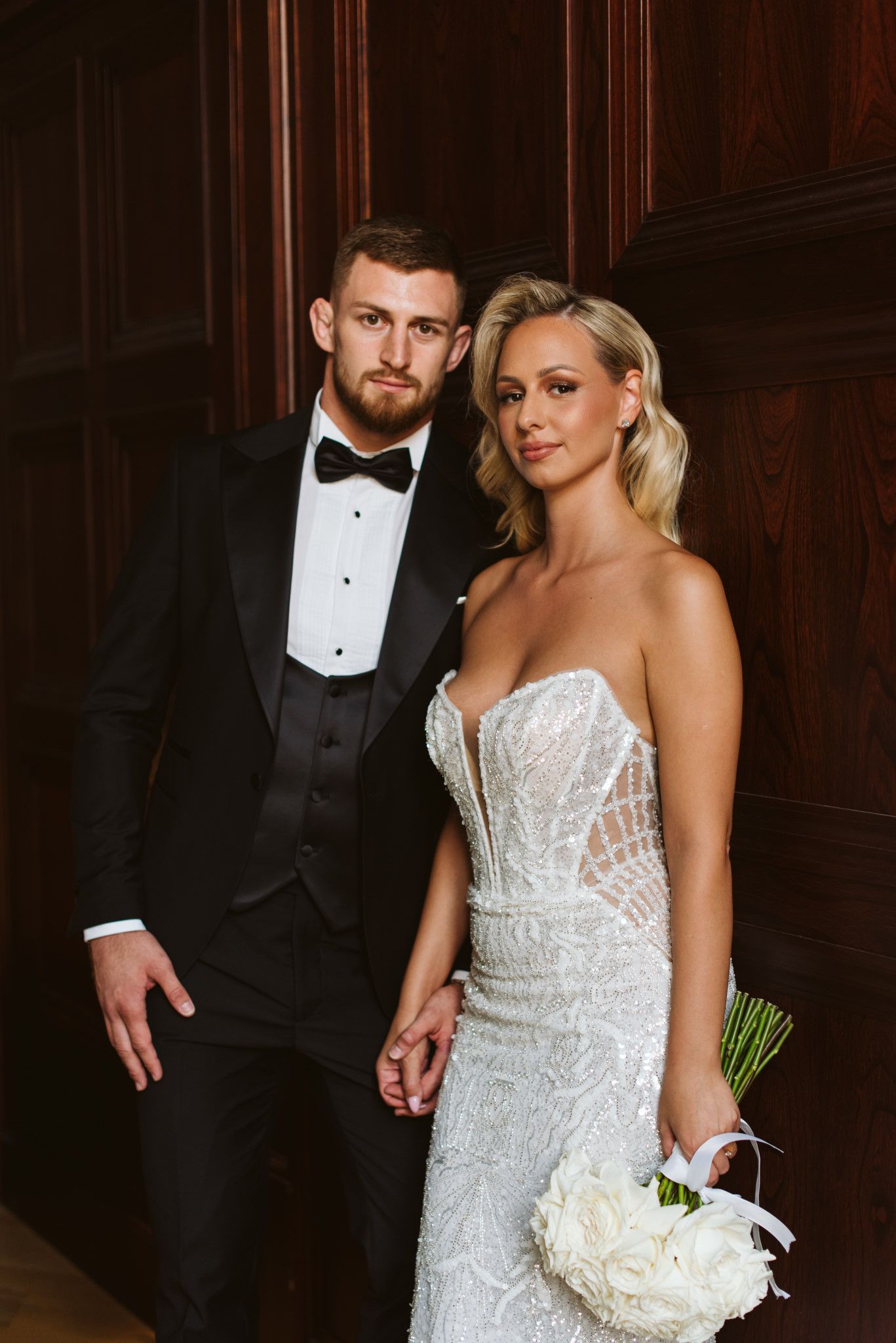 A bride holds a white bouquet while posing with her groom in formal wedding attire against wood paneled walls.
