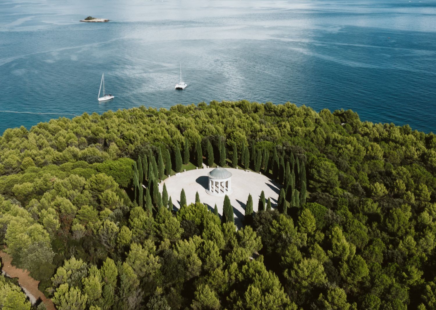 Aerial view of a white building surrounded by lush forest on a coastal peninsula with boats in the blue ocean.  Istria Wedding