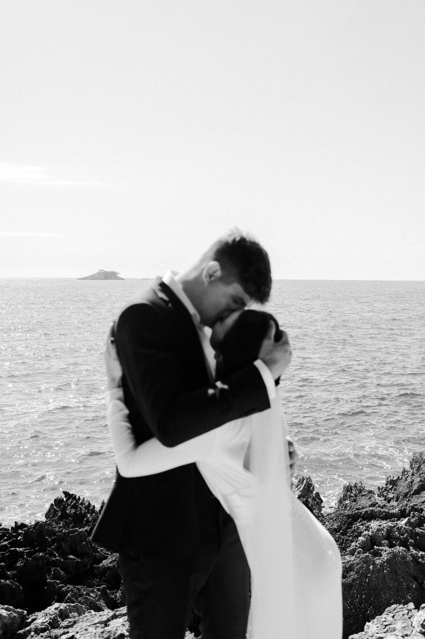 A romantic black and white photo of a couple embracing by the ocean with rocky cliffs in the background.