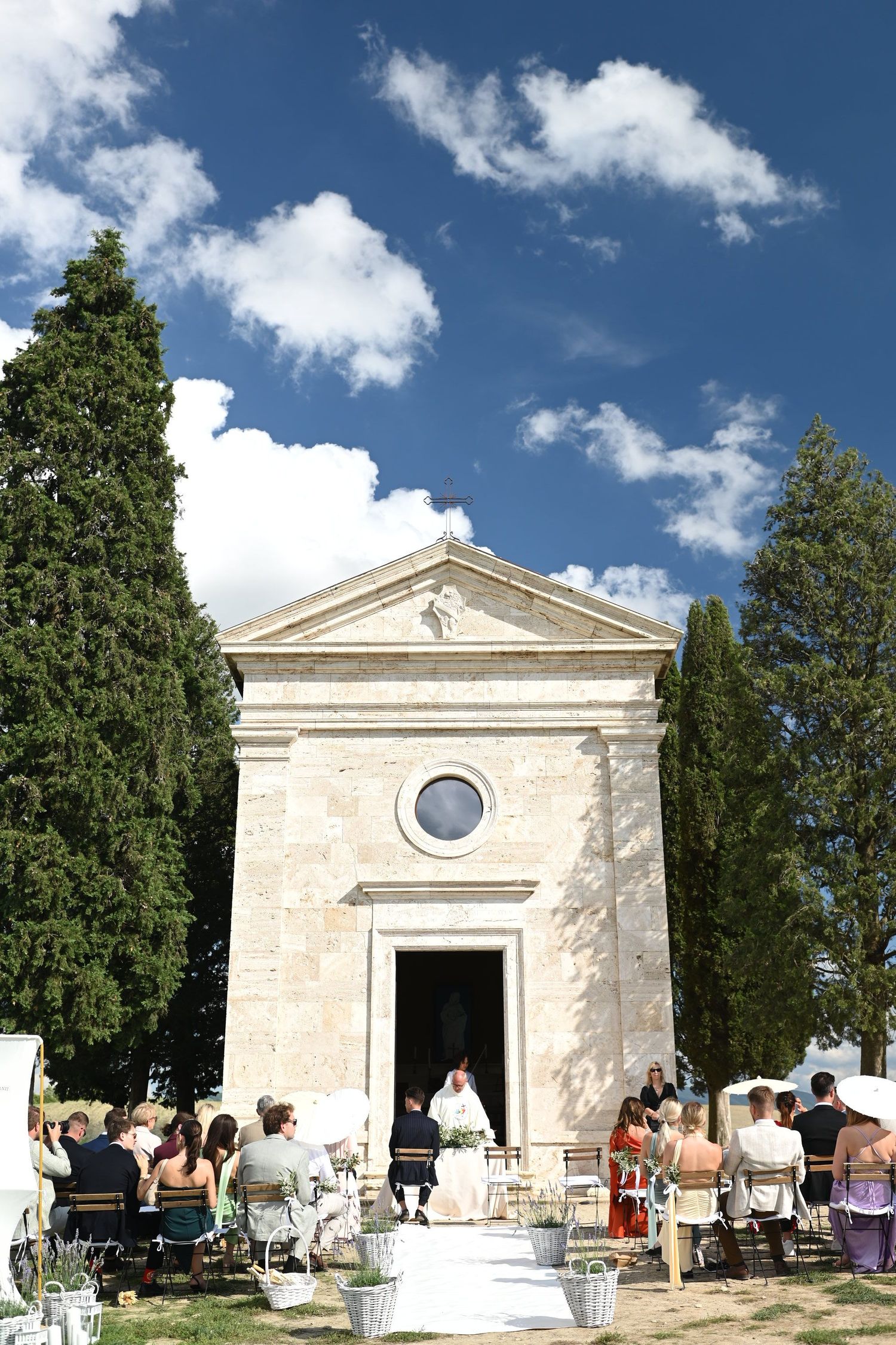 A small stone chapel with cypress trees hosts an outdoor wedding ceremony under blue skies in Tuscany.