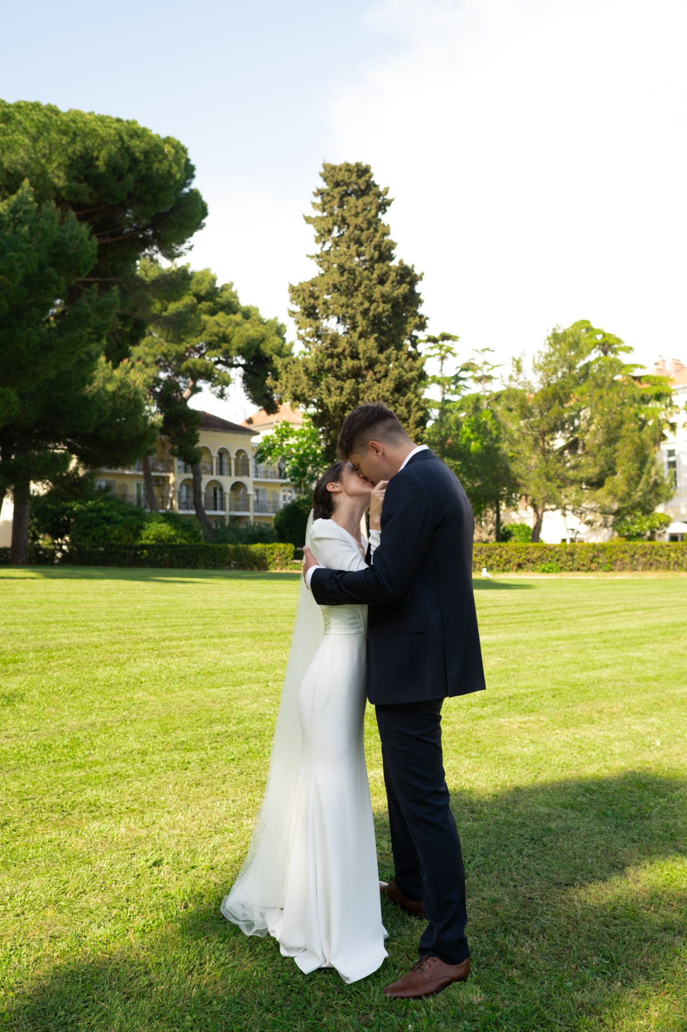 A romantic wedding moment captured on a lush green lawn with a mansion and tall trees in the background.