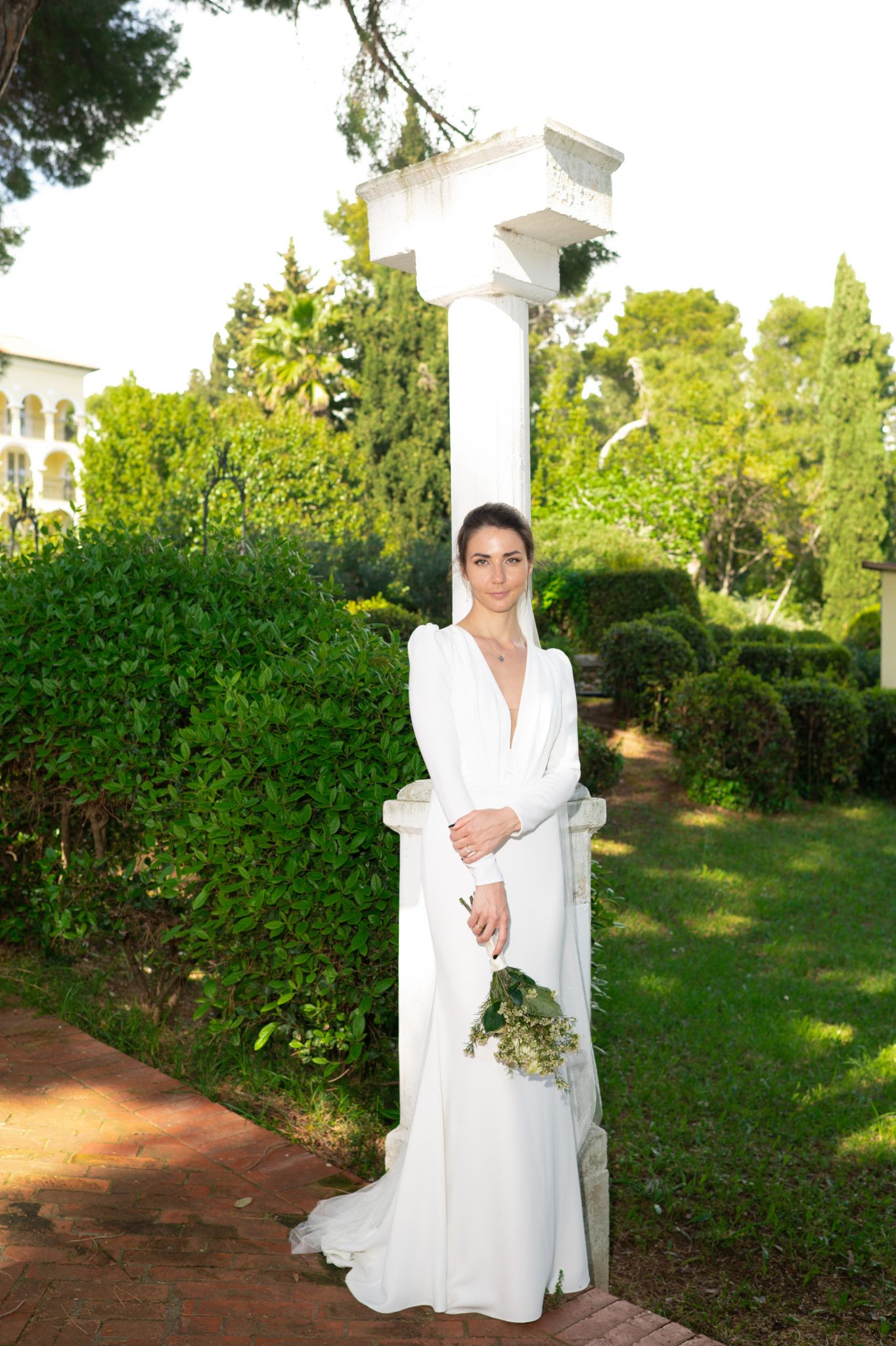 A bride in a flowing white gown poses in a garden setting with classical architecture.