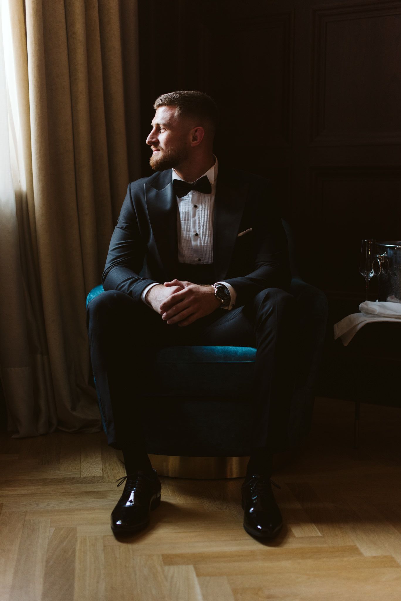 Groom in formal attire seated in teal armchair with moody lighting.