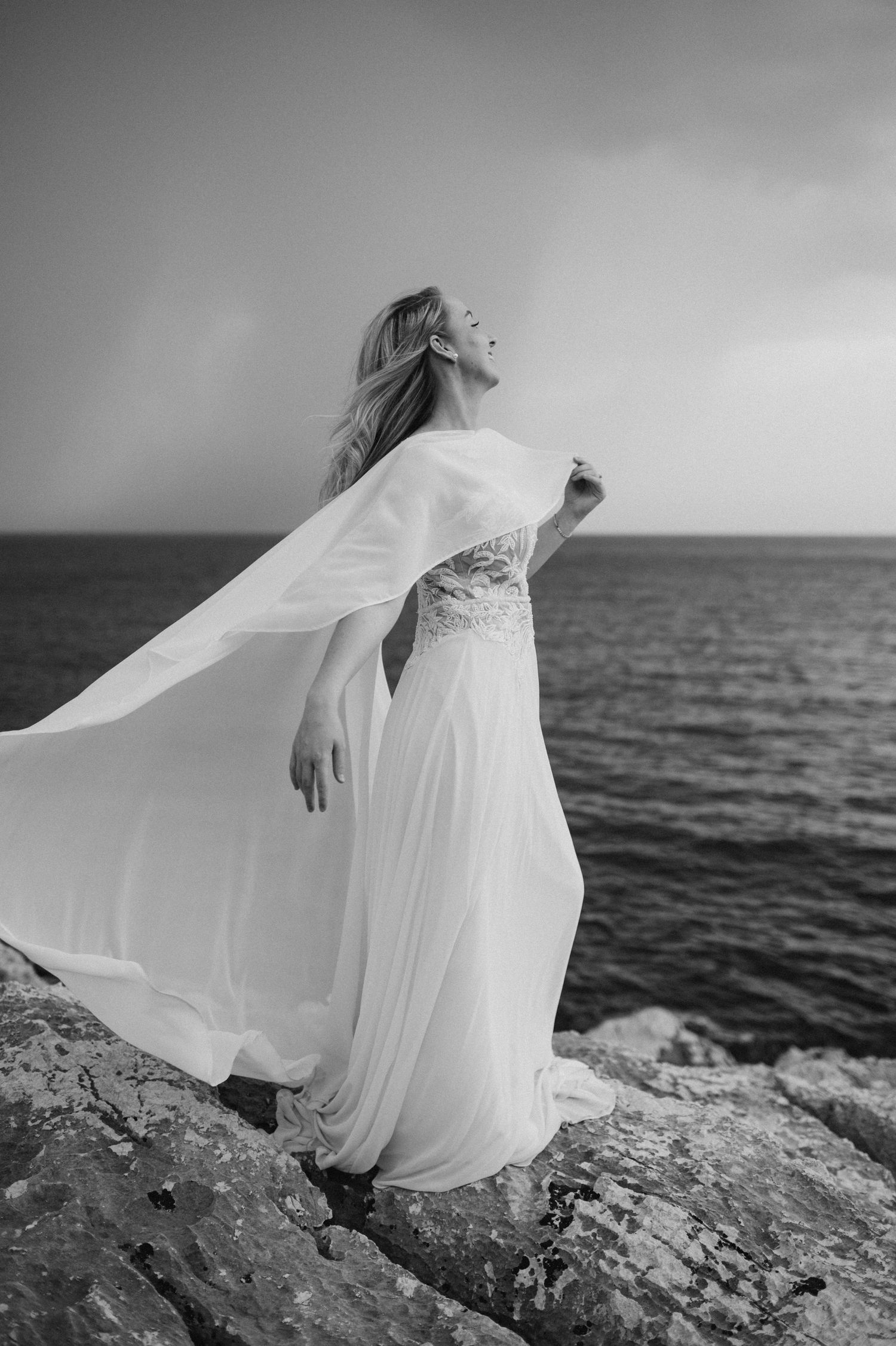 A flowing white dress billows in the wind against a dramatic black and white coastal landscape with ocean waves.