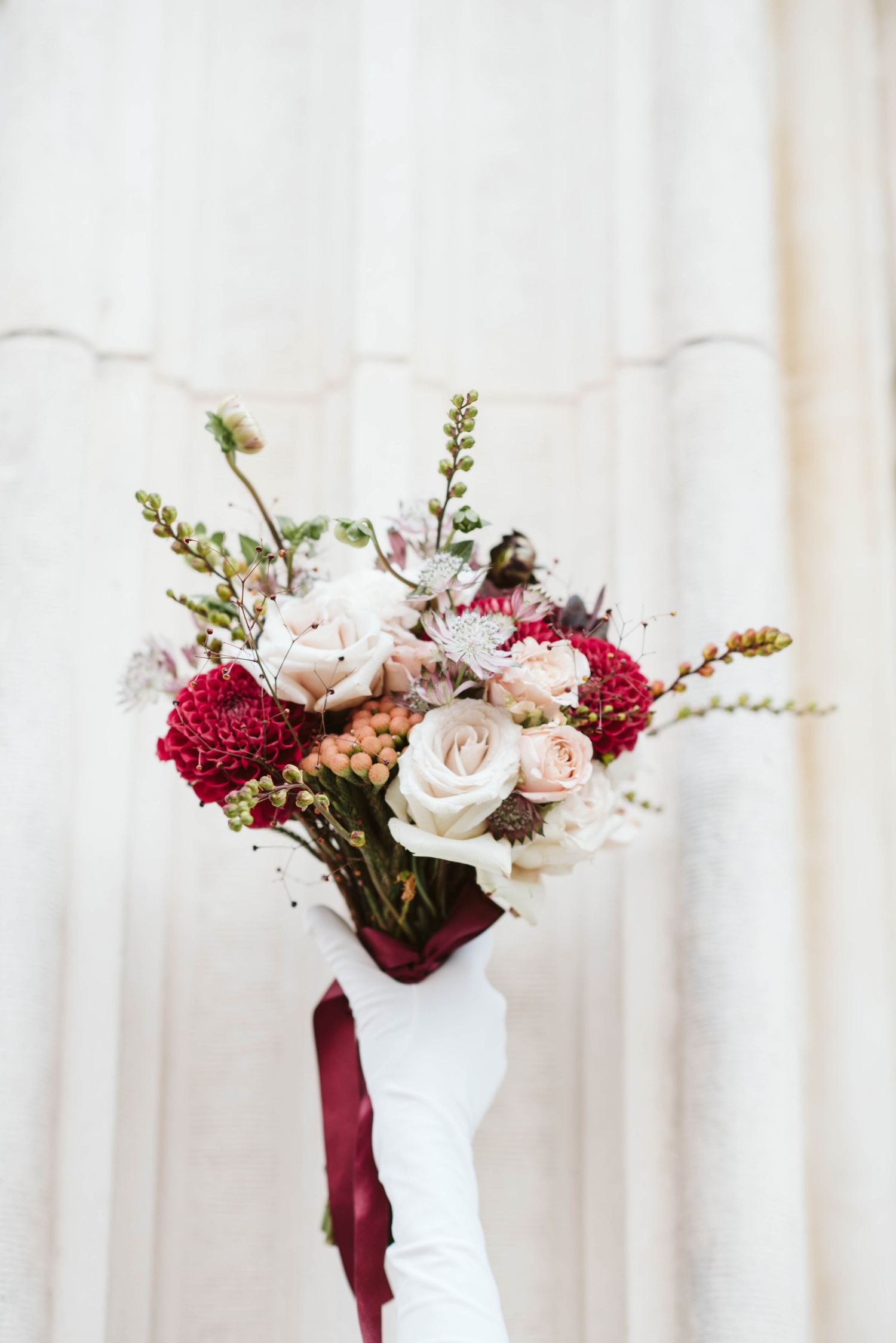 Three elegant white vases with burgundy and blush floral arrangements displayed against a light curtained background.