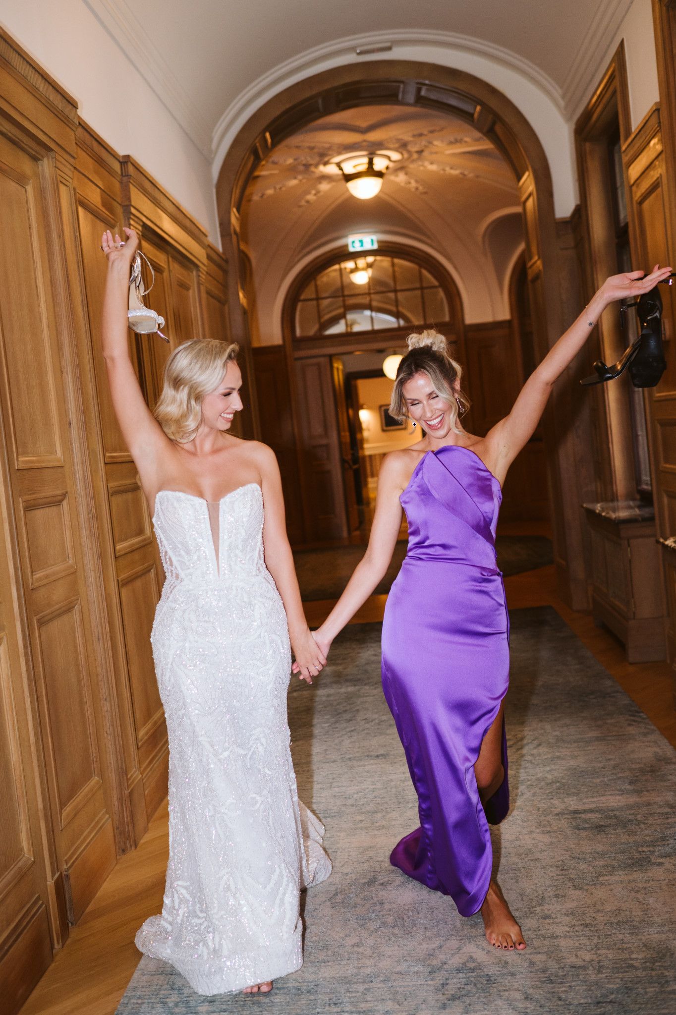 Two women in white and purple dresses dancing joyfully in grand hallway.