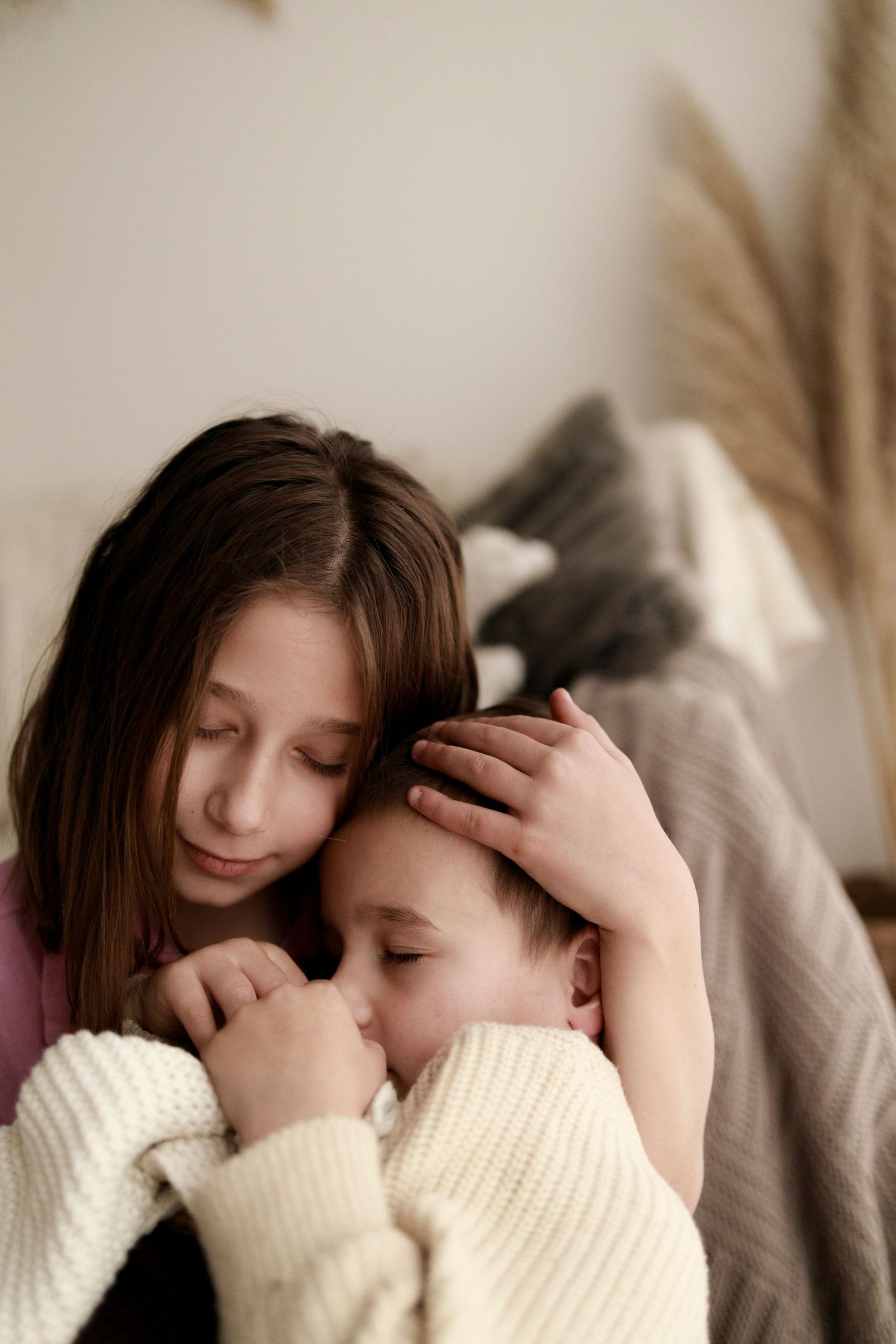a girl sitting down in front of pampas grass with eyes closed whilst cradling a little boy who has his eyes closed in soft light