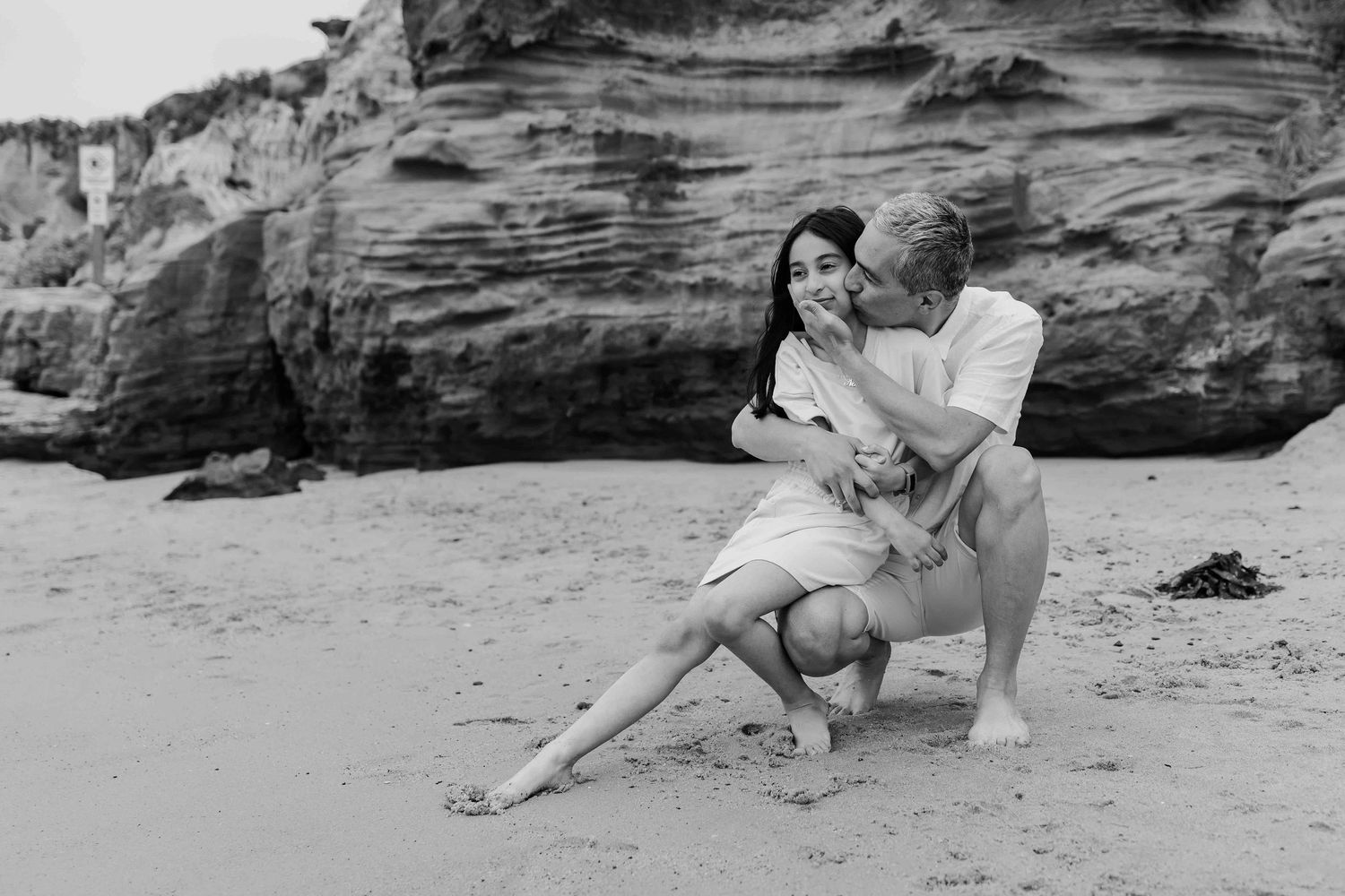 a black and white image of a young girl looking out and sitting on man's knee as he holds and kisses her face whilst kneeling on sand