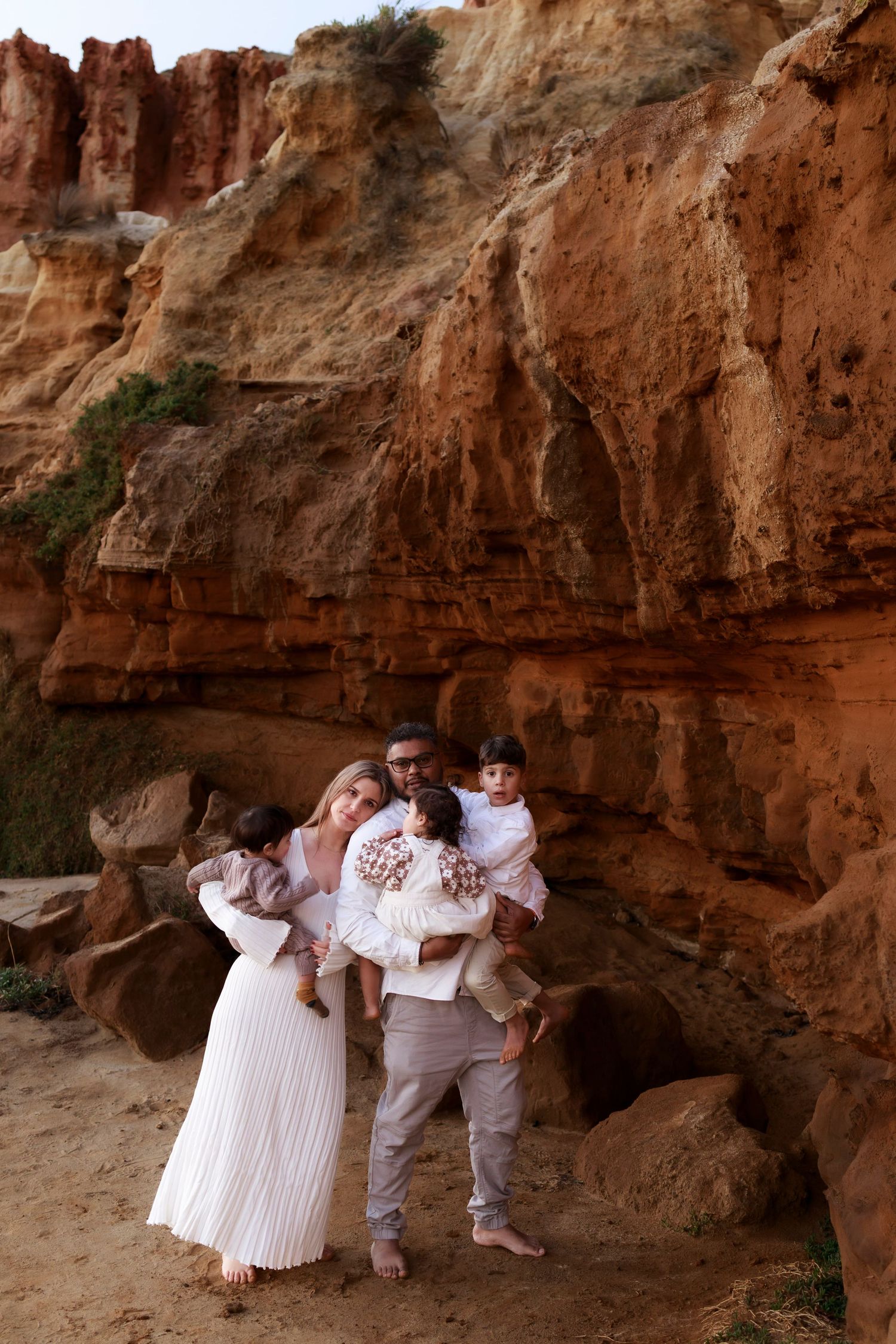 a man and woman standing cuddling together wearing neutral colours whilst holding 3 young children next to a rock cliff side and sand