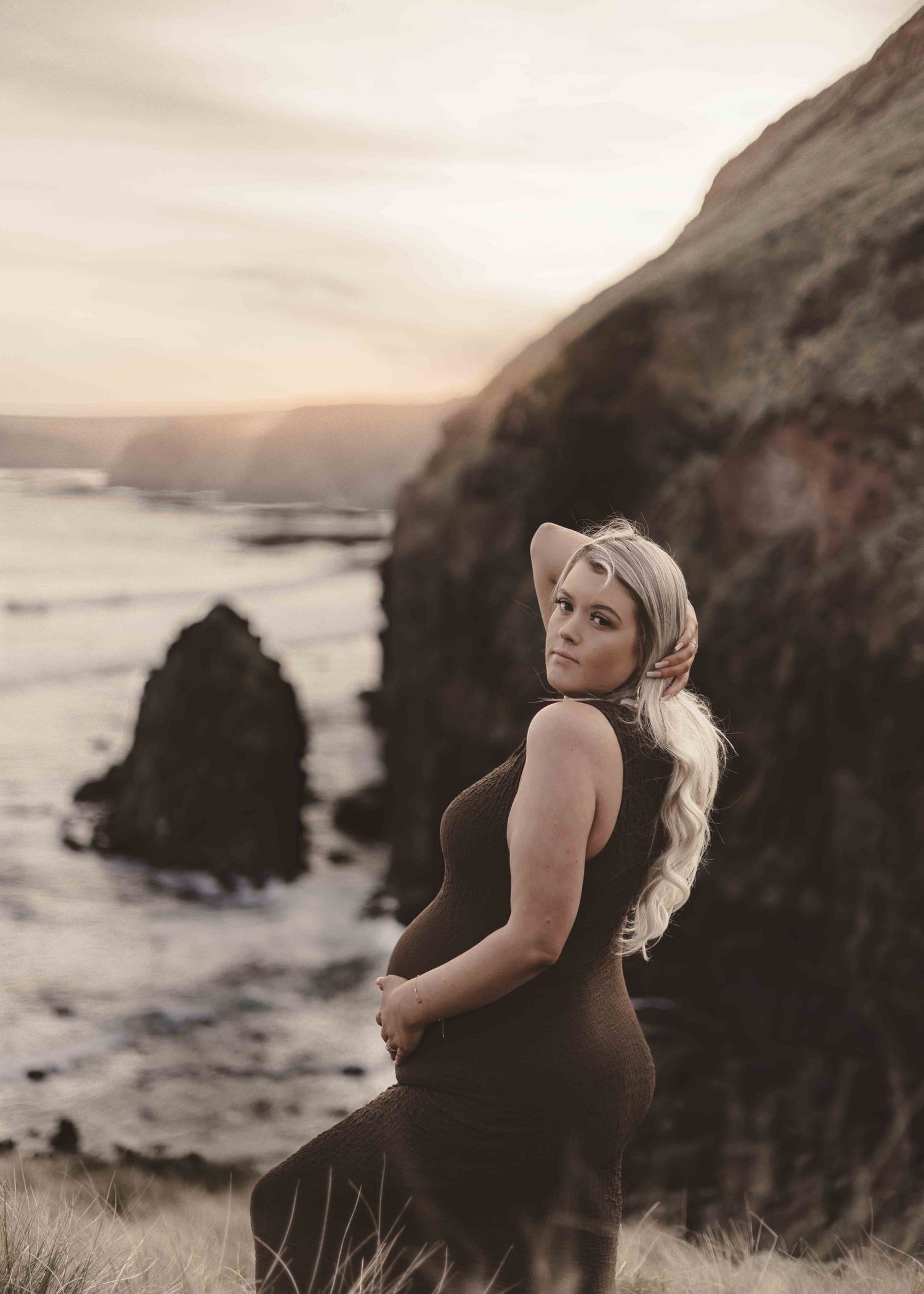 a blonde pregnant woman standing overlooking a cliffside wearing a brown dress whilst looking directly into the camera over her shoulder