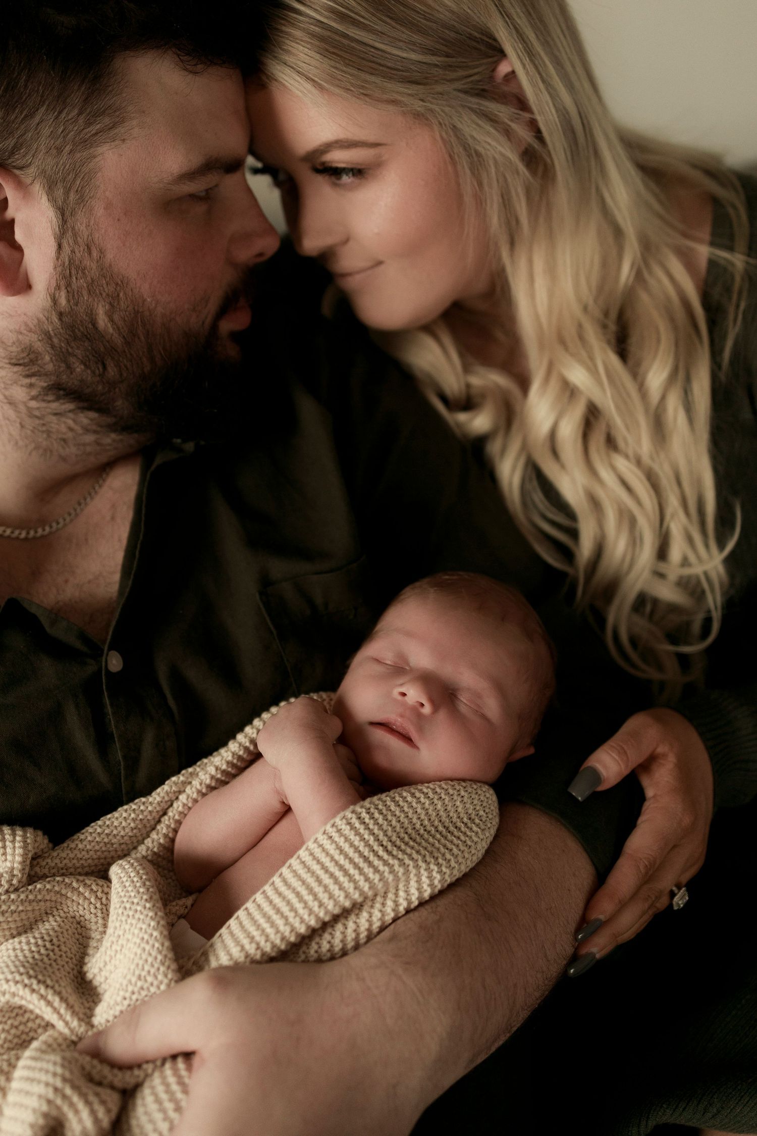 a newborn baby sleeping in a knitted beige blanket with arms showing and being held by mother and father as they look at each other smiling