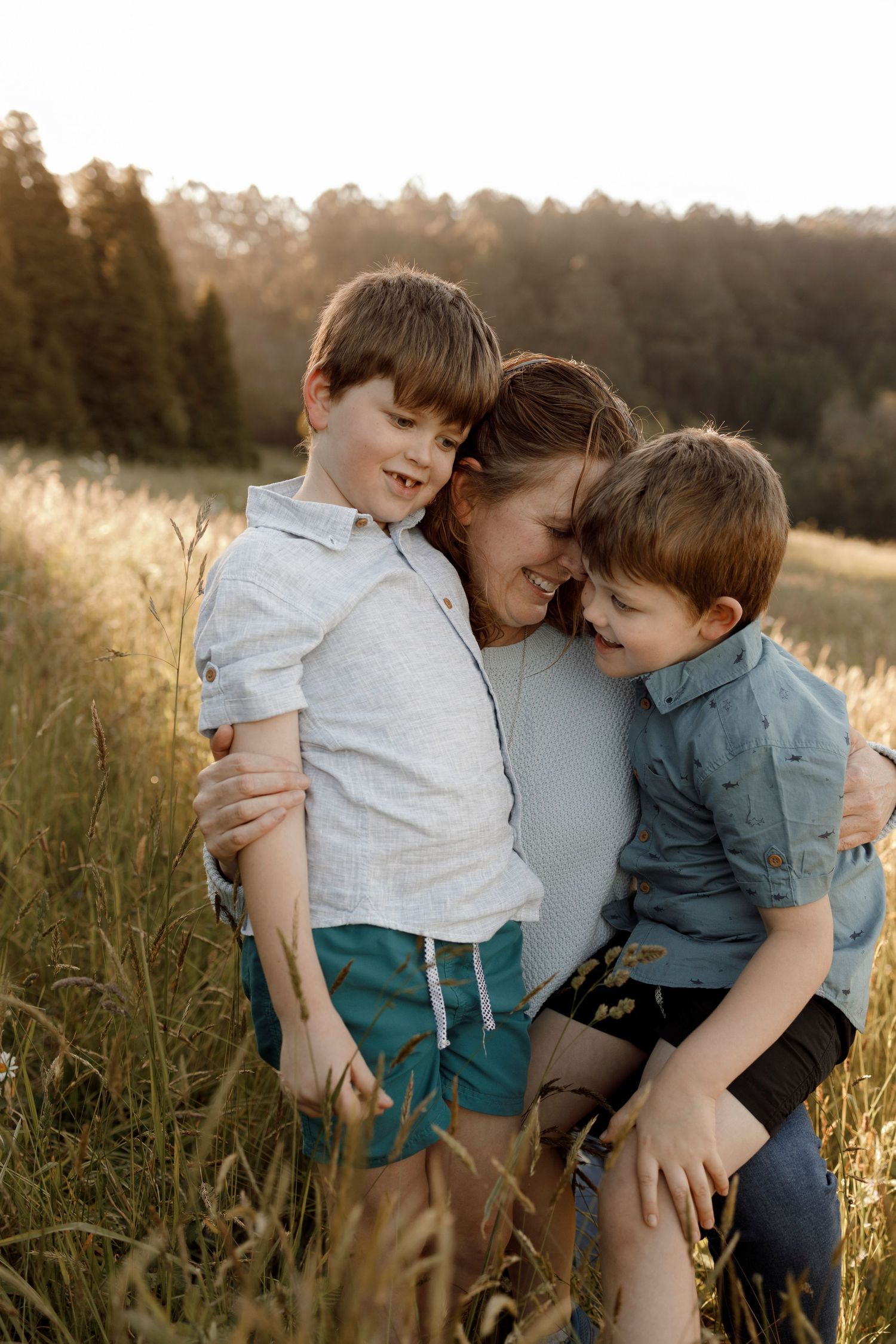 a woman wearing a blue jumper and jeans holding kneeling down smiling in a field of grass with two young boys cuddling into her
