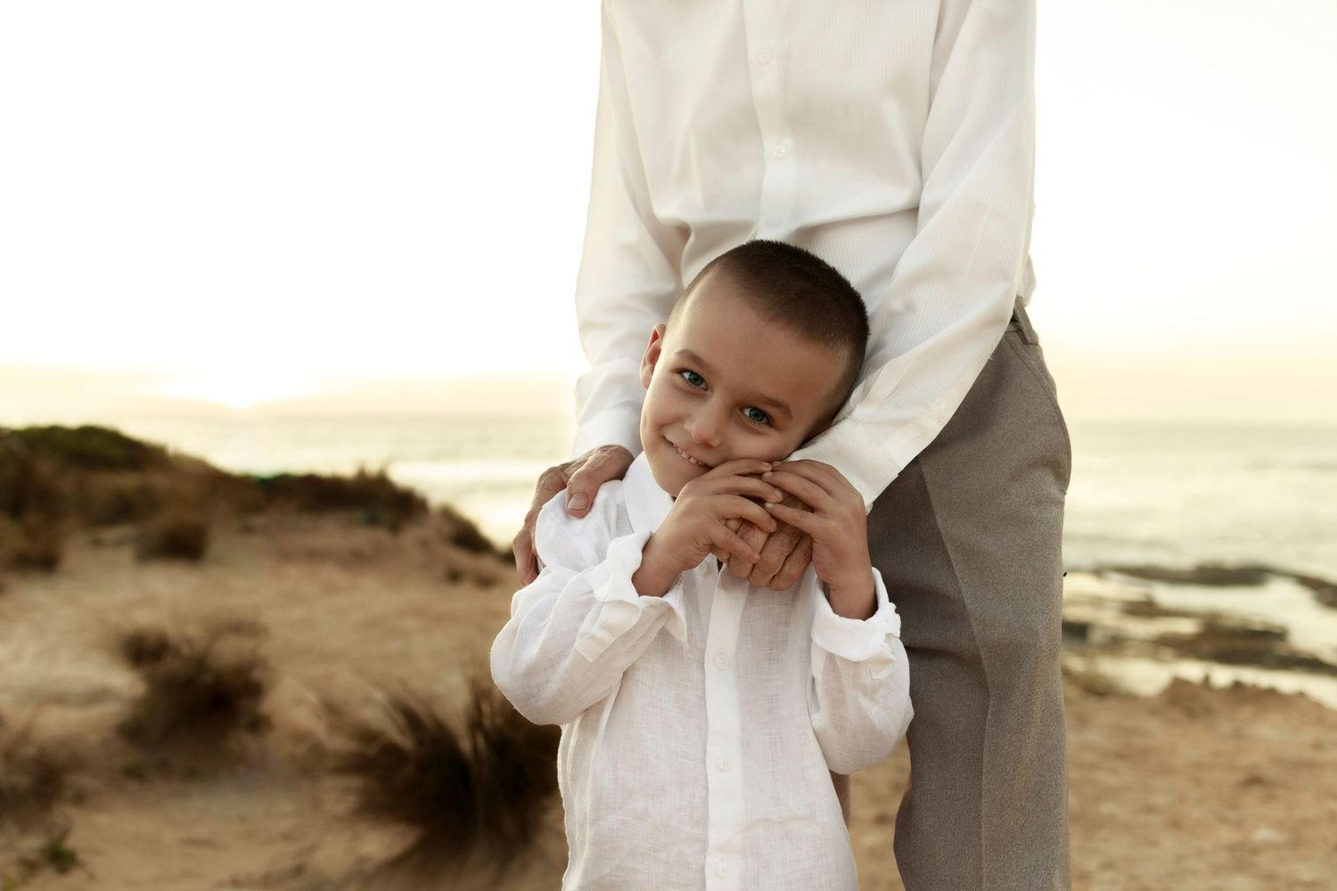 a little boy wearing a white shirt smiling looking into the camera in front of an elderly man who has his hand under the boys face