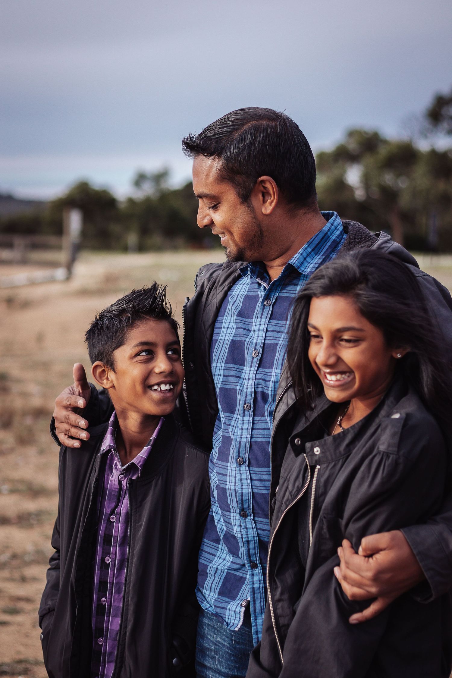 a man wearing a blue checked shirt looking down at young boy and arm around young girl standing on sand wearing a black leather jacket