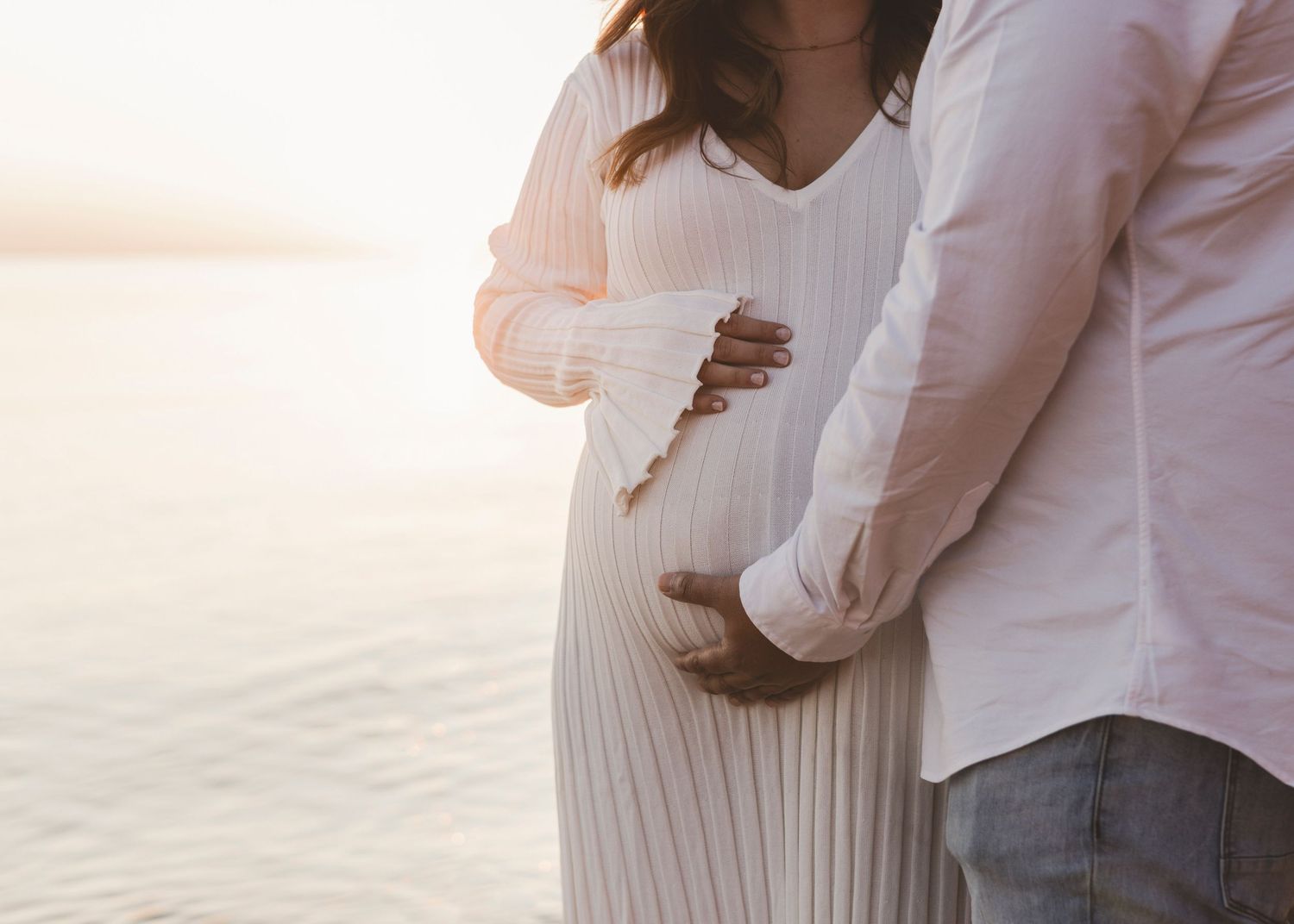a pregnant woman standing in front of the water at the beach with a man's hand placed underneath holding her belly