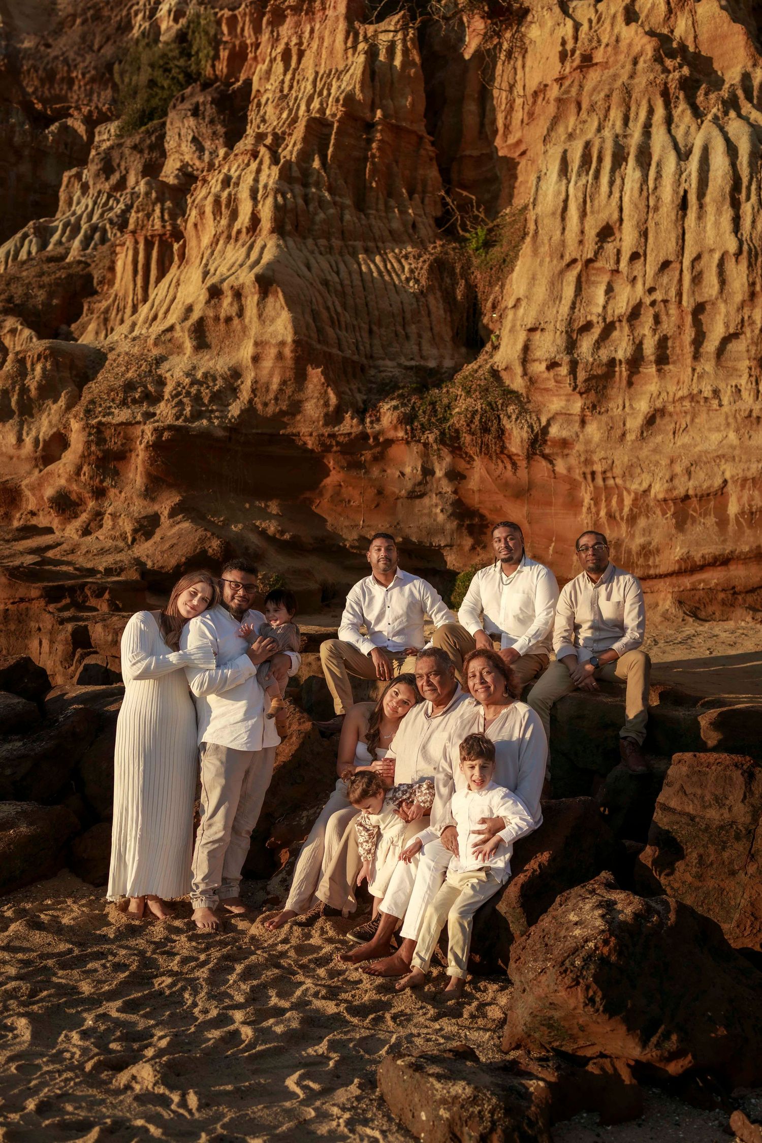 a large family standing together wearing neutral colours whilst cuddling close and smiling in the sunset by cliff side and rocks