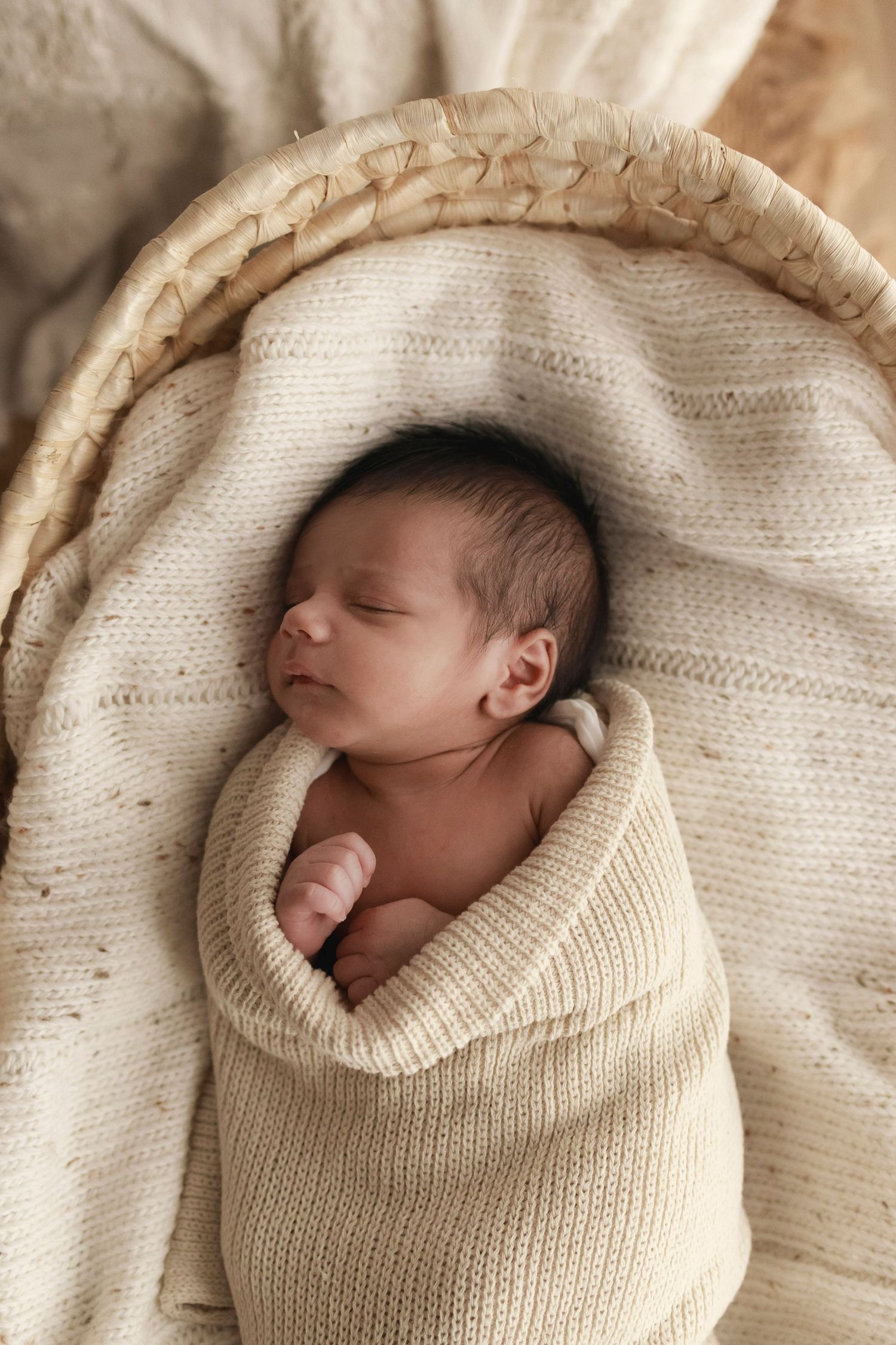 a newborn baby sleeping in a moses basket on top of a neutral coloured blanket wrapped up in a beige knitted blanket with hands showing