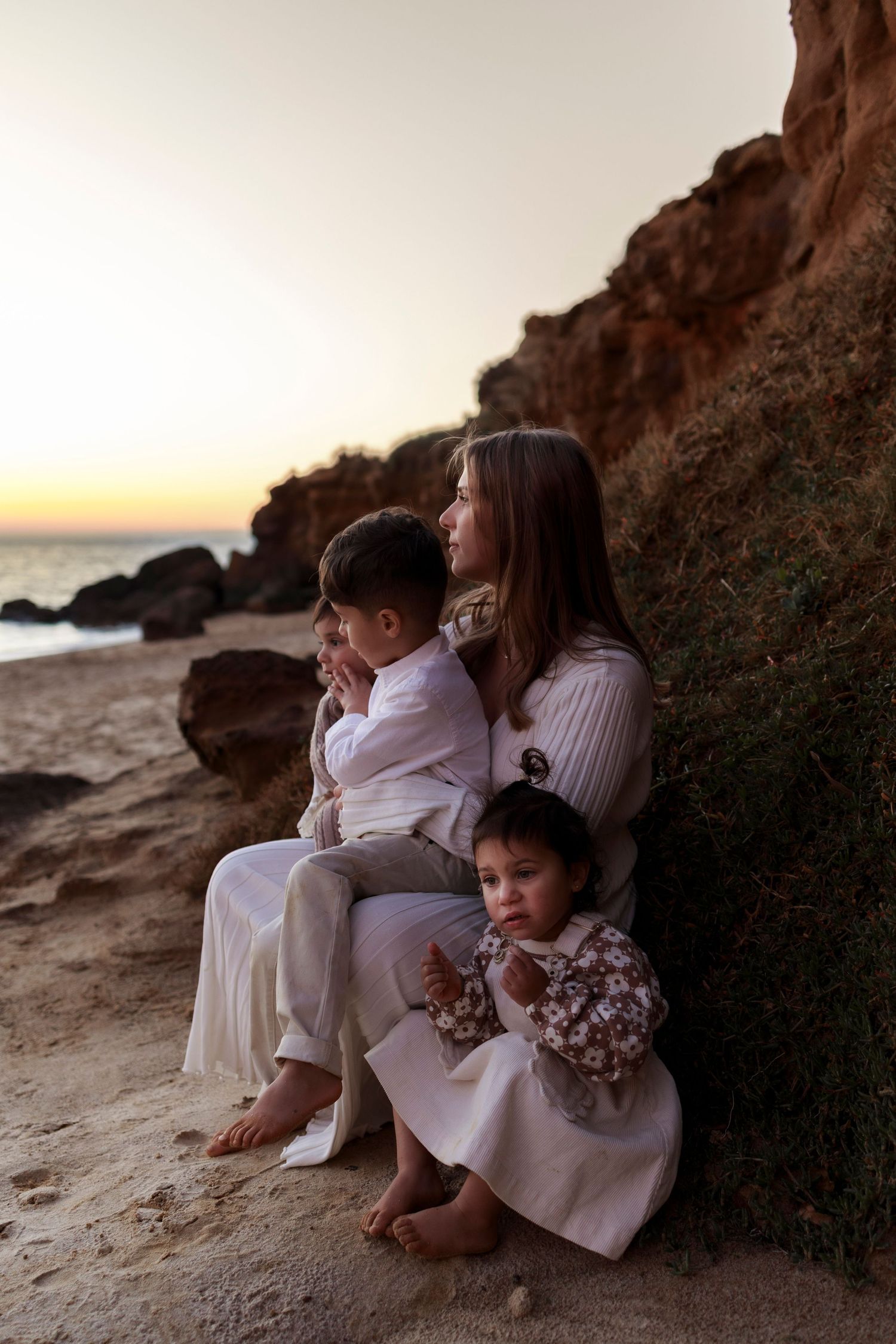 a lady wearing a white dress kneeling down against a rock on sand holding a baby and two older children in sunset light