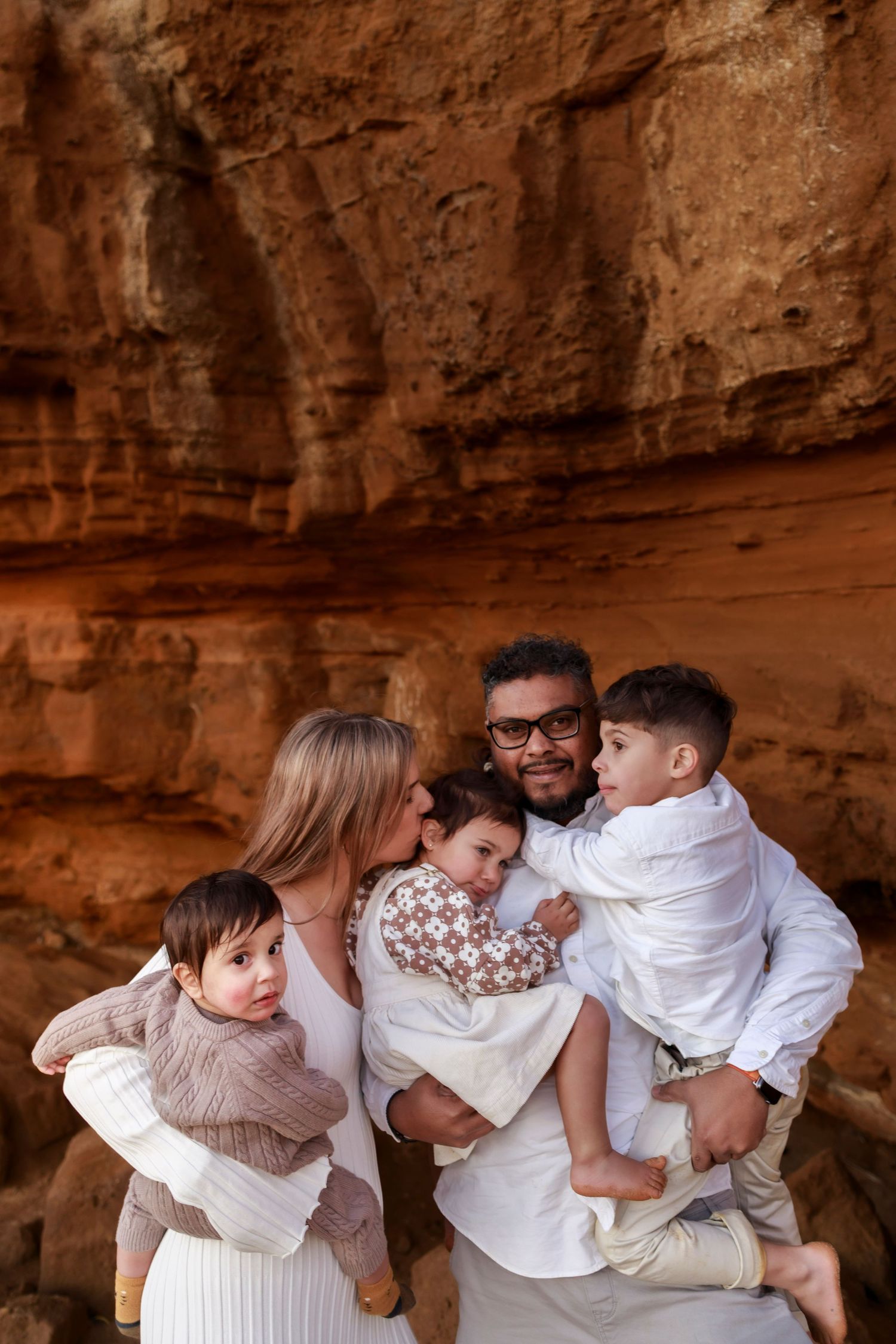 a man a man and woman standing cuddling together wearing neutral colours whilst holding 3 young children next to a rock cliff side and sand