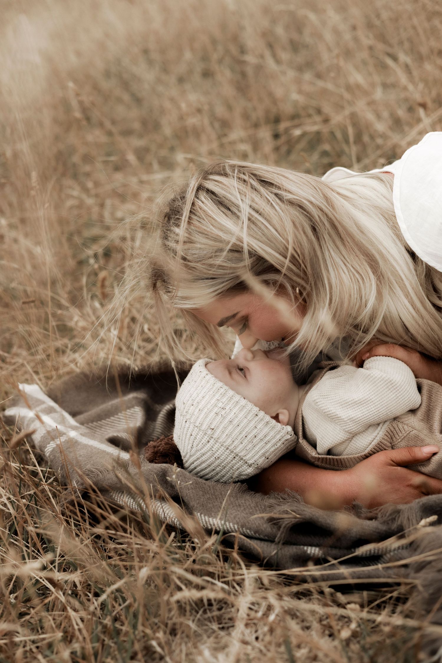 a baby wearing a beanie laying on top of a rug in grass lovingly looking up at a woman's smiling face with blonde hair