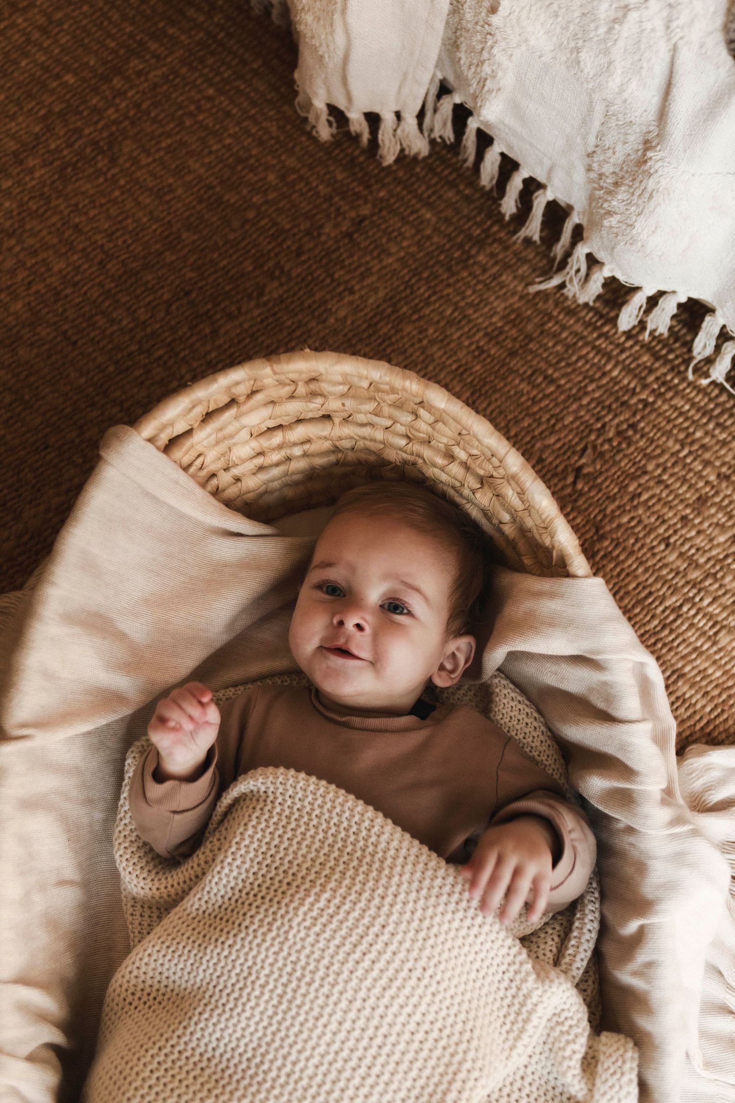 a happy baby laying in a moses basket looking up wrapped in a beige knitted blanket with soft tones
