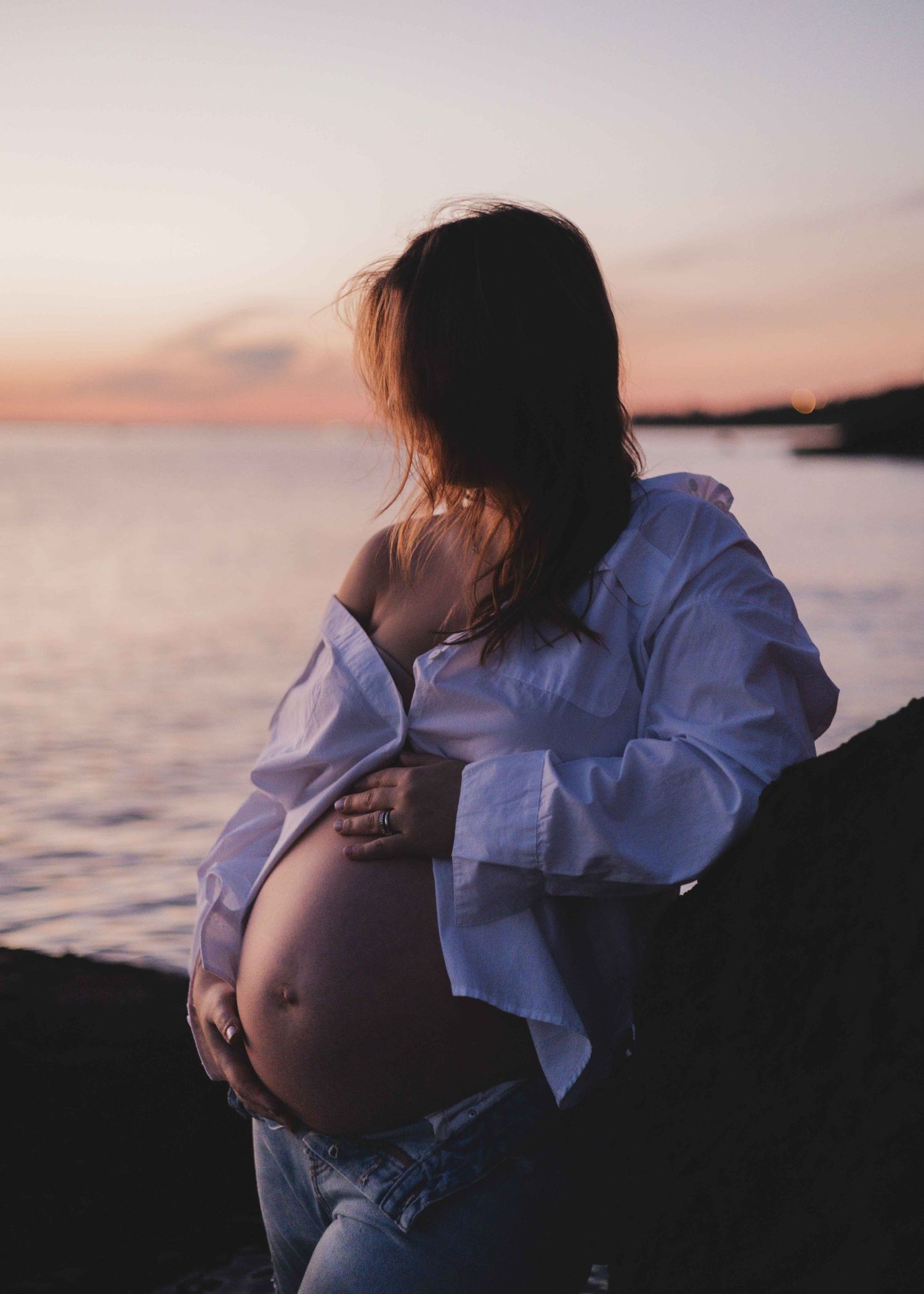 a pregnant woman standing leaning against a rock at a beach with open white shirt and holding her belly with her hair covering her face