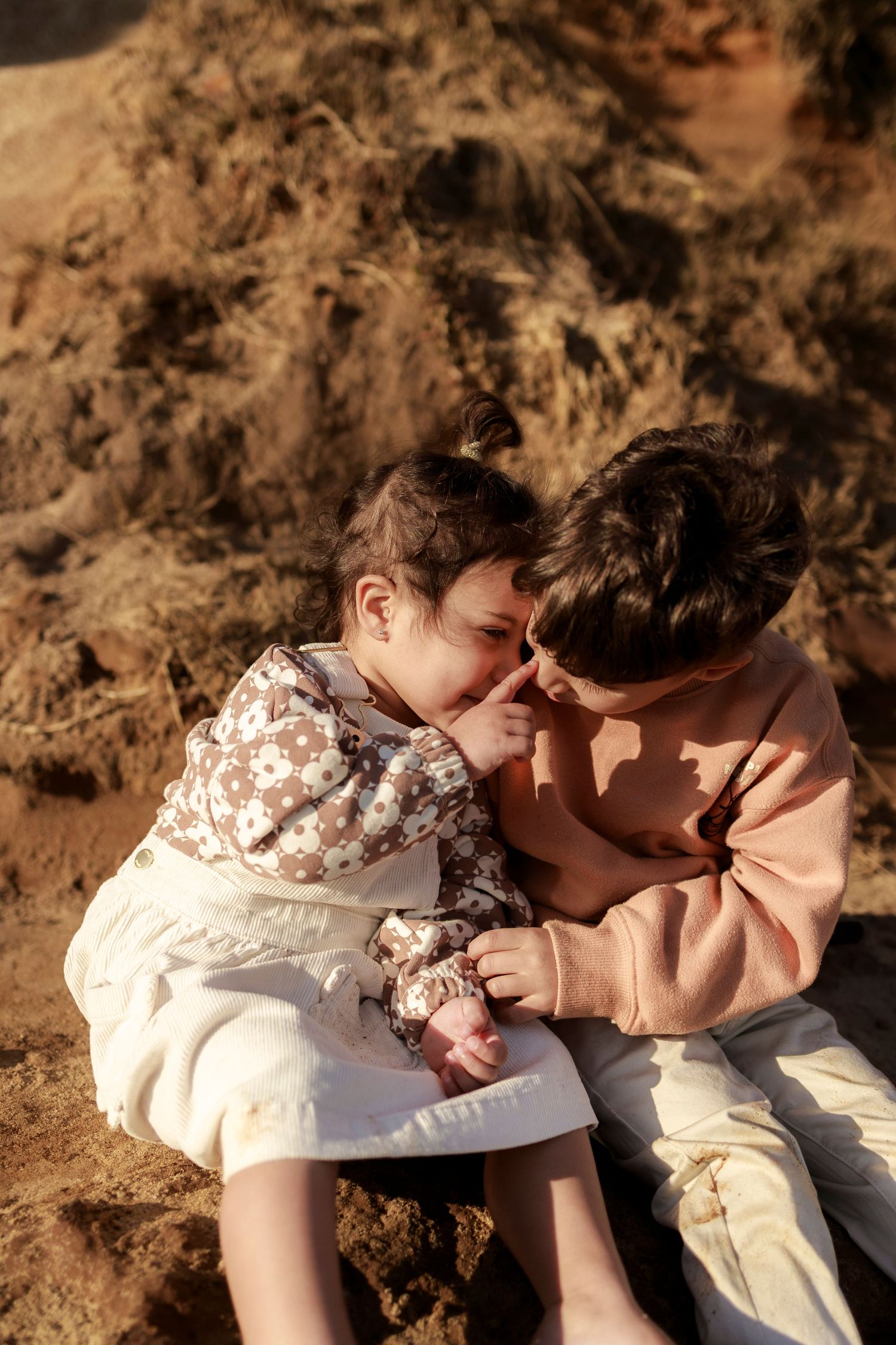 a young girl and boy sit together on rocks in sunset light being playful as the girl touches the boys nose