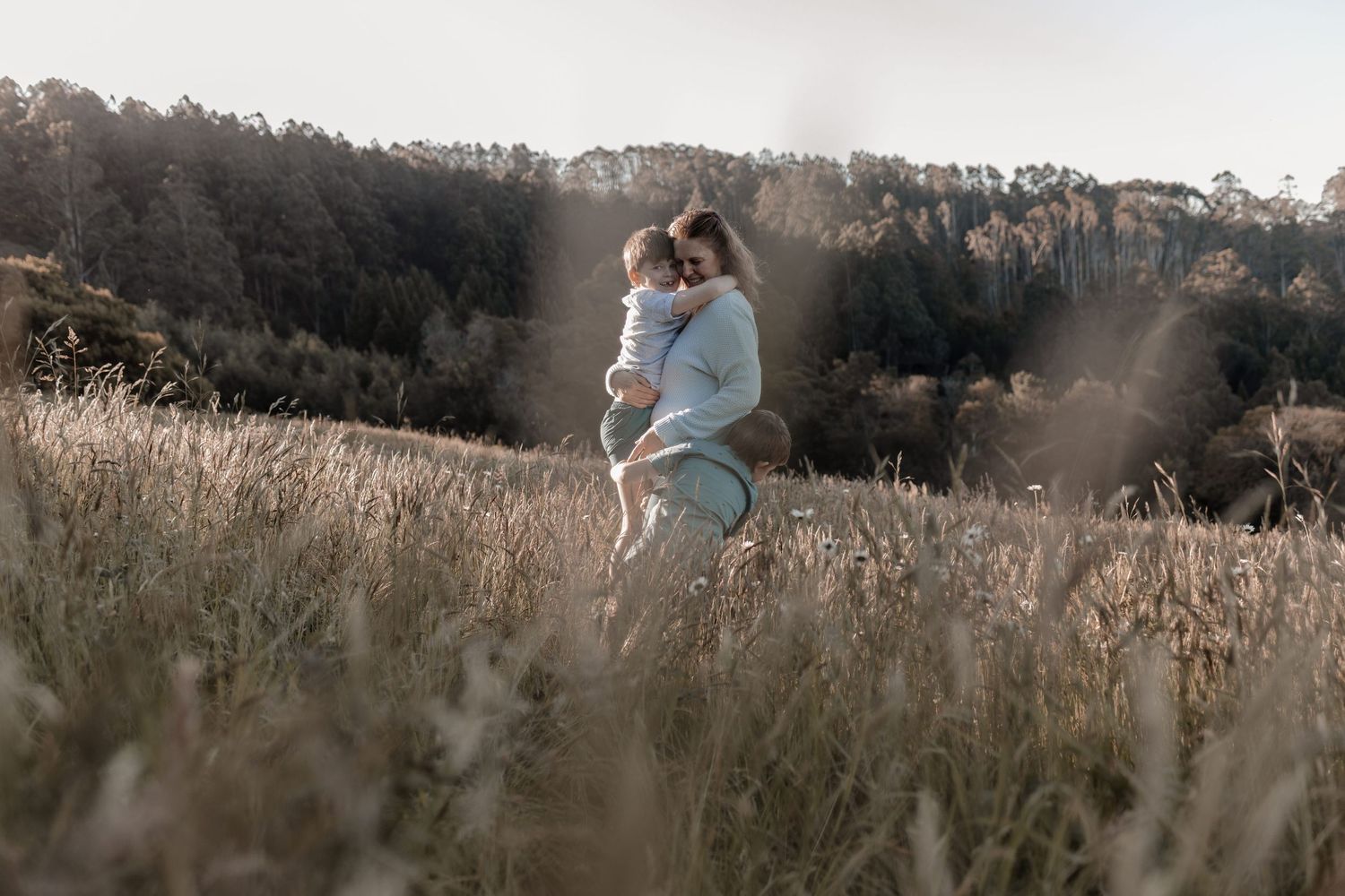a woman wearing a blue jumper and jeans holding a young boy in a field of grass whilst  another young boy hugs her legs